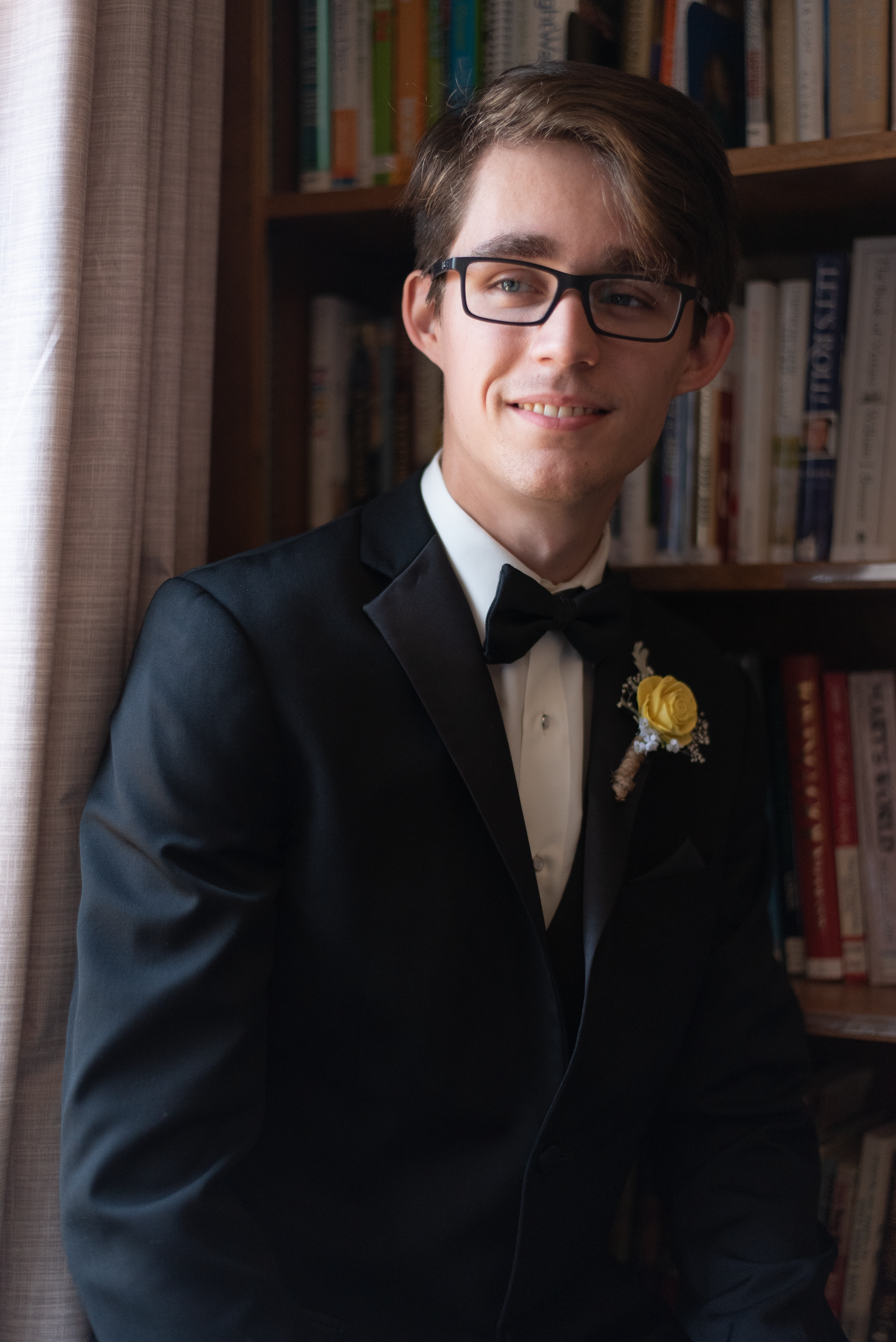 Dapper groom in black tuxedo with yellow boutonniere smiling in front of a bookshelf.