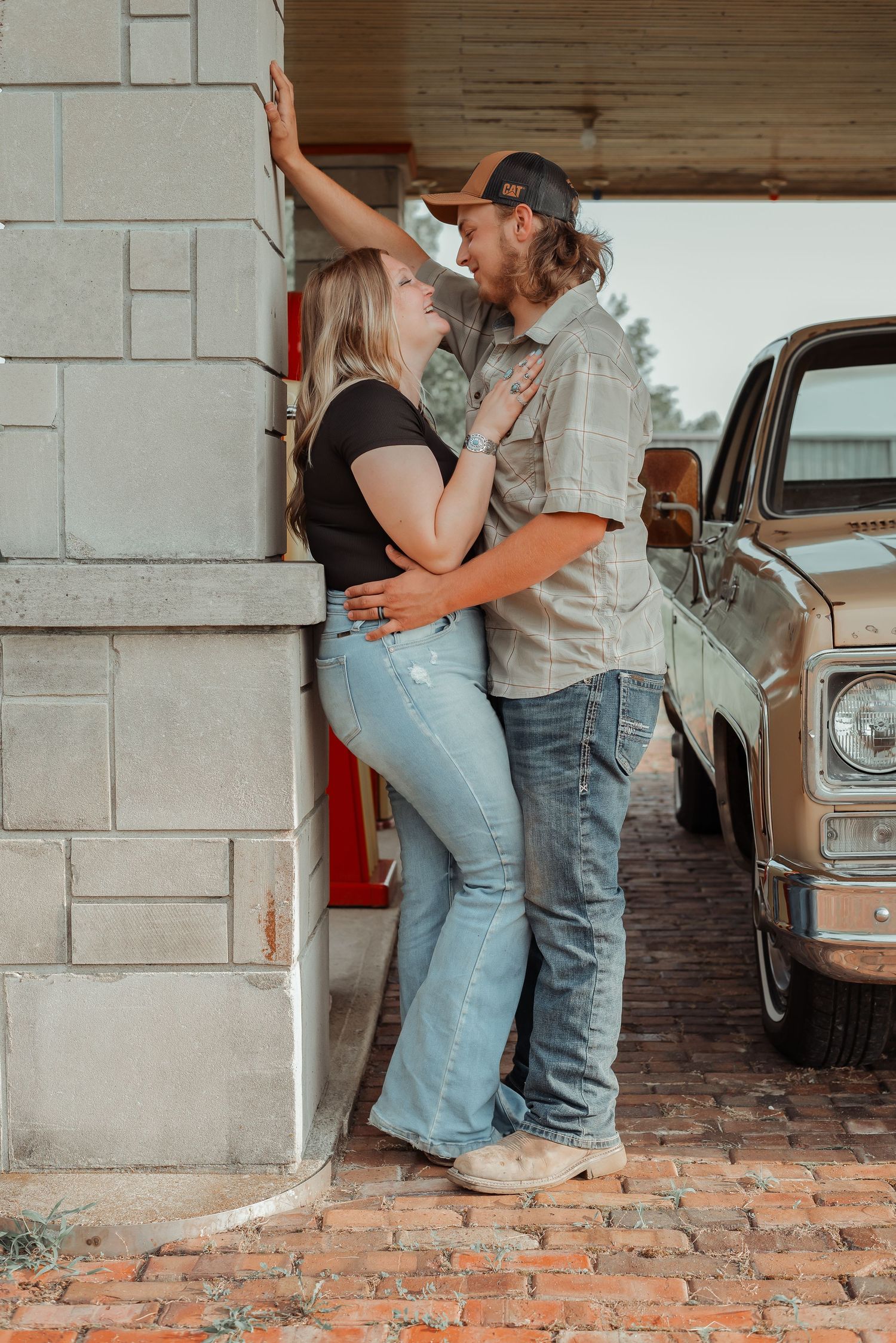 Couple in casual denim outfits share a tender moment leaning against a brick pillar near a vintage truck.