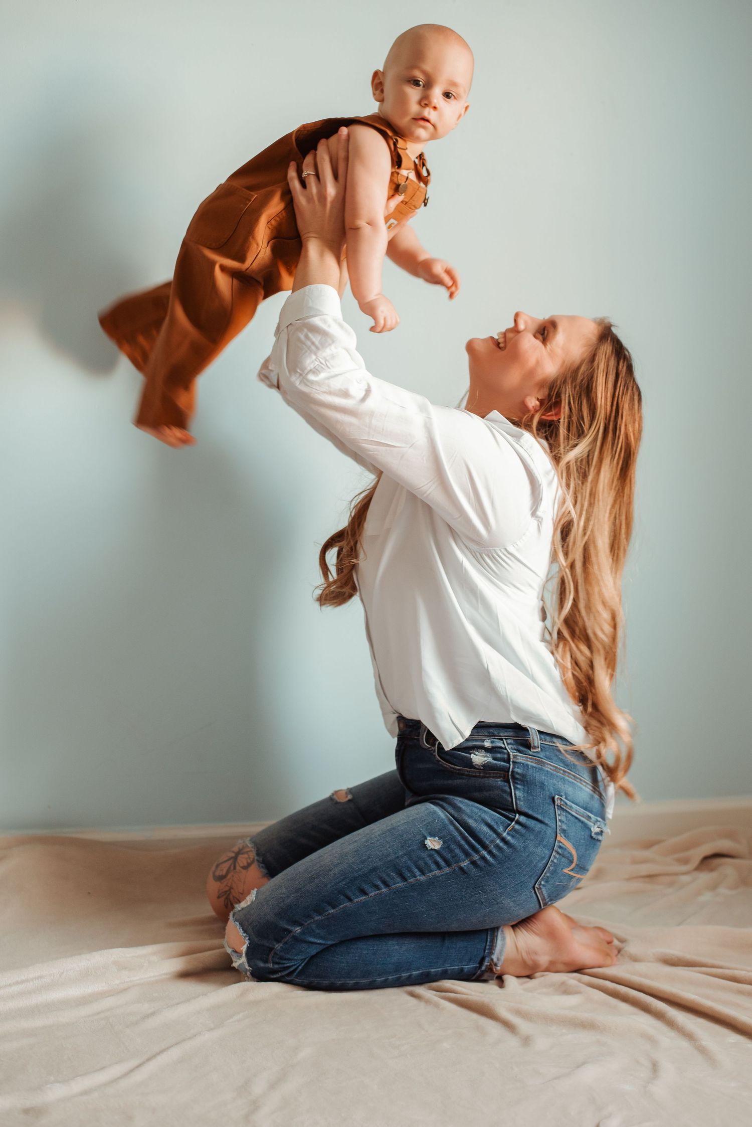 A joyful mother kneels on a bed, lifting her baby in a rust dress overhead against a light blue wall.