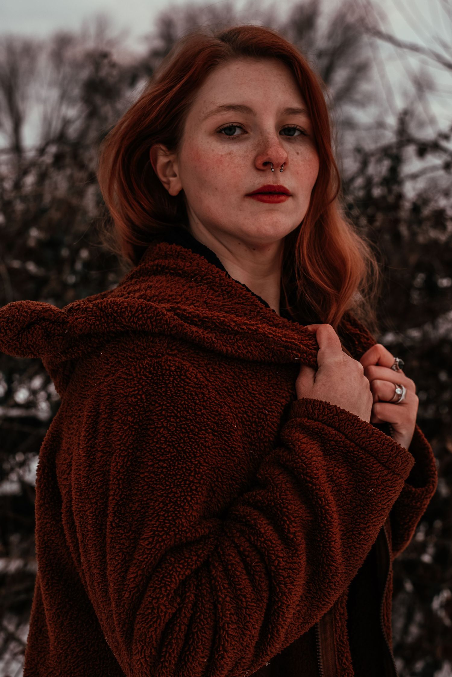 Close-up portrait of a freckled redhead woman in a brown fleece hoodie coat outdoors.