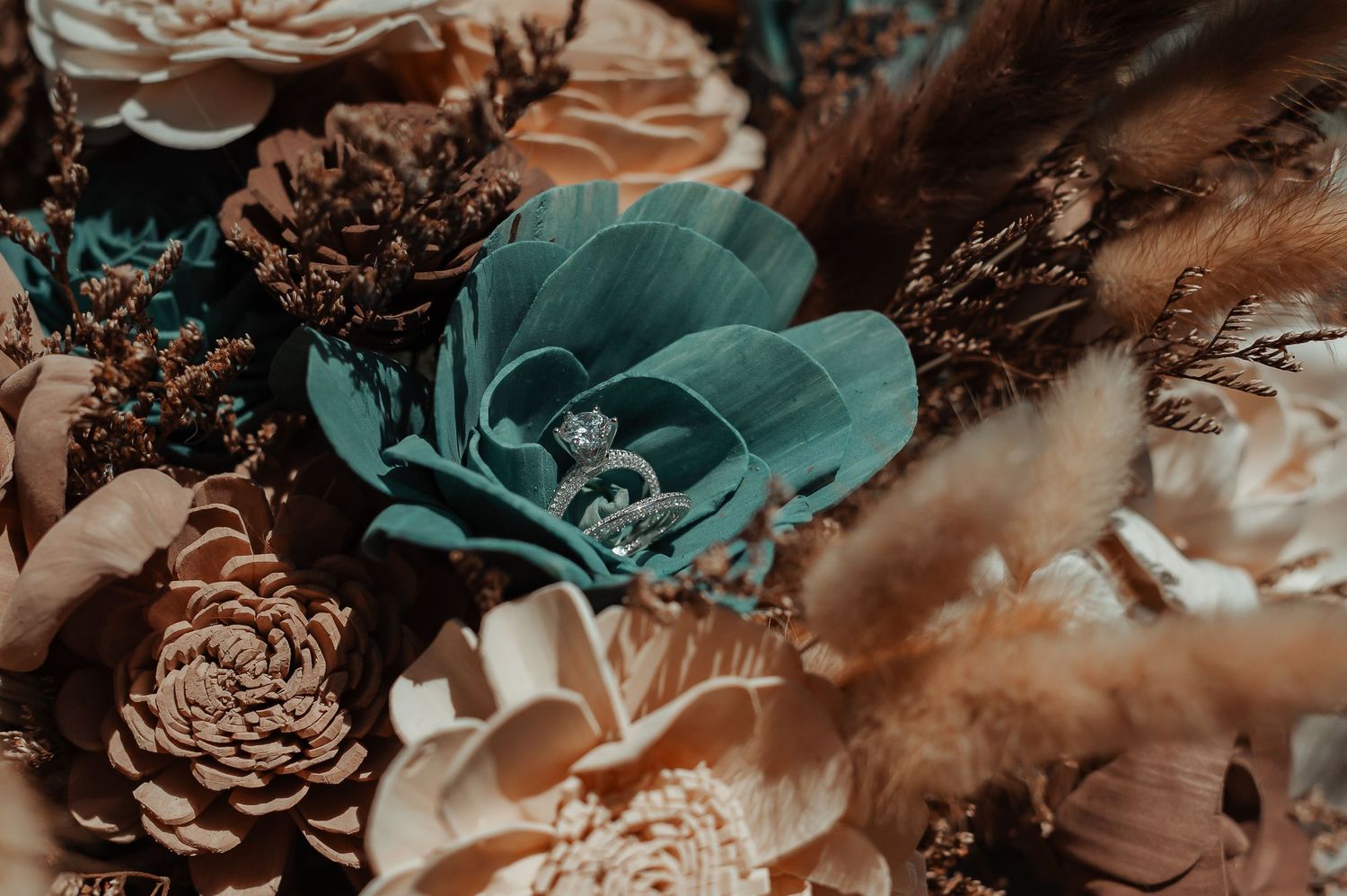 Close-up of a diamond engagement ring nestled among teal and blush dried wedding bouquet flowers.