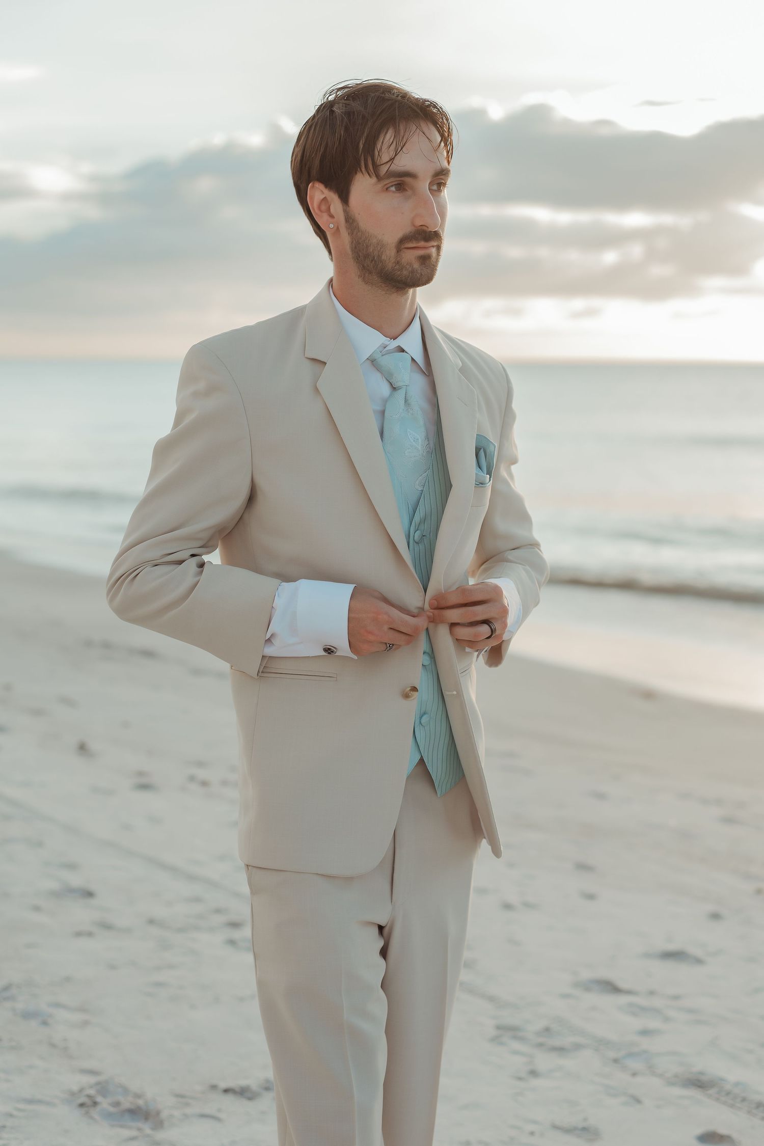 Handsome groom in tan suit with mint tie and vest standing on beach at sunset.