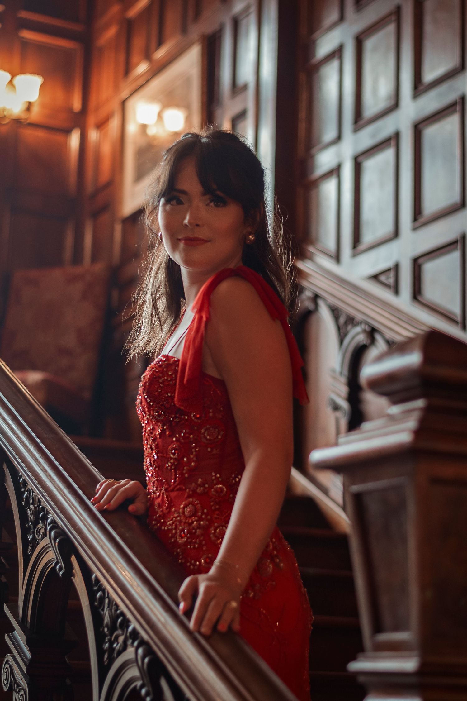 Woman in red beaded gown posing on ornate wooden staircase in a classic elegant interior.