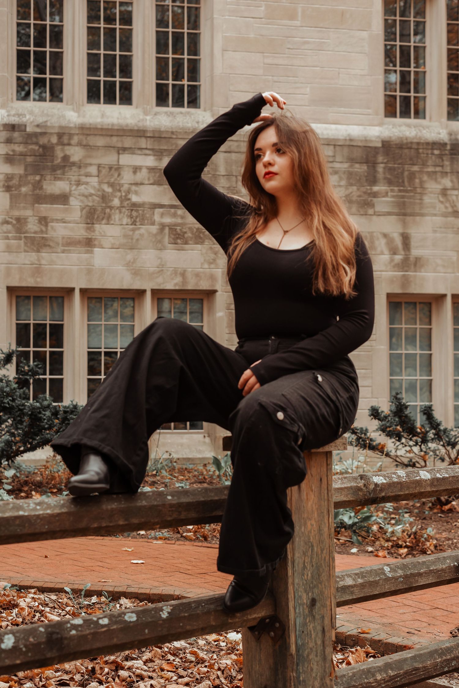 Young woman in black bodysuit and wide-leg pants poses on wooden fence near stone building.