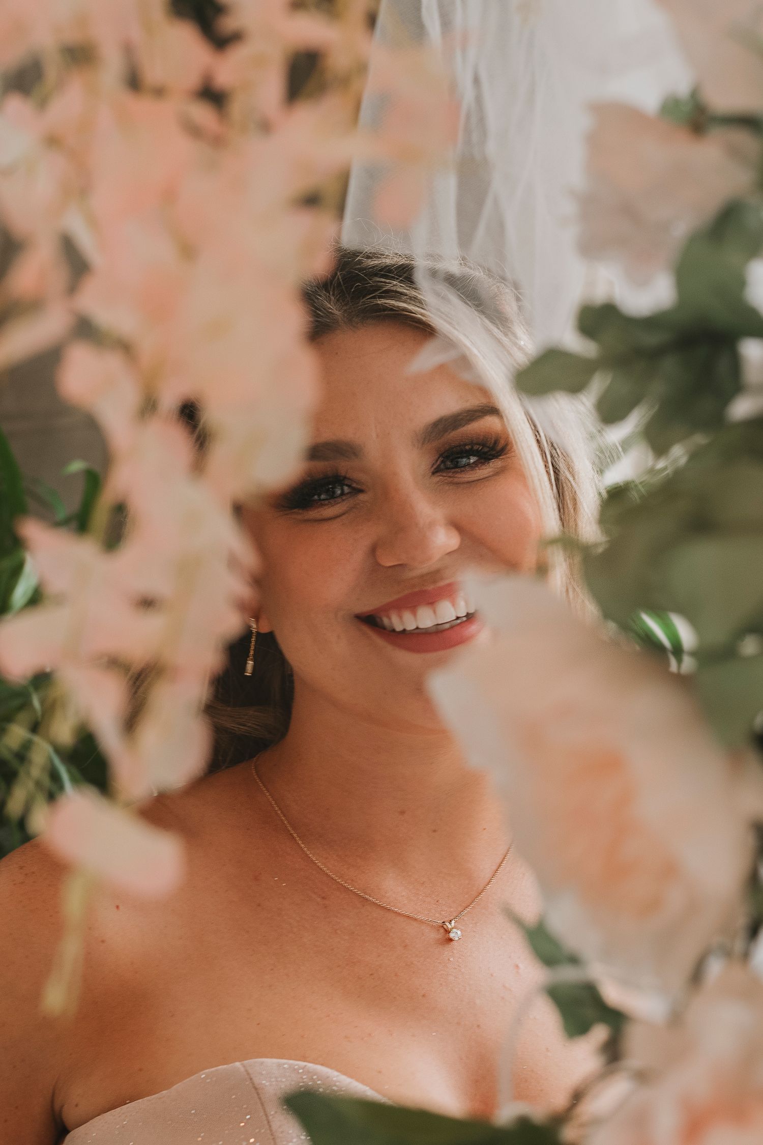Smiling bride in veil framed by soft pink blossoms and greenery wearing a delicate necklace.