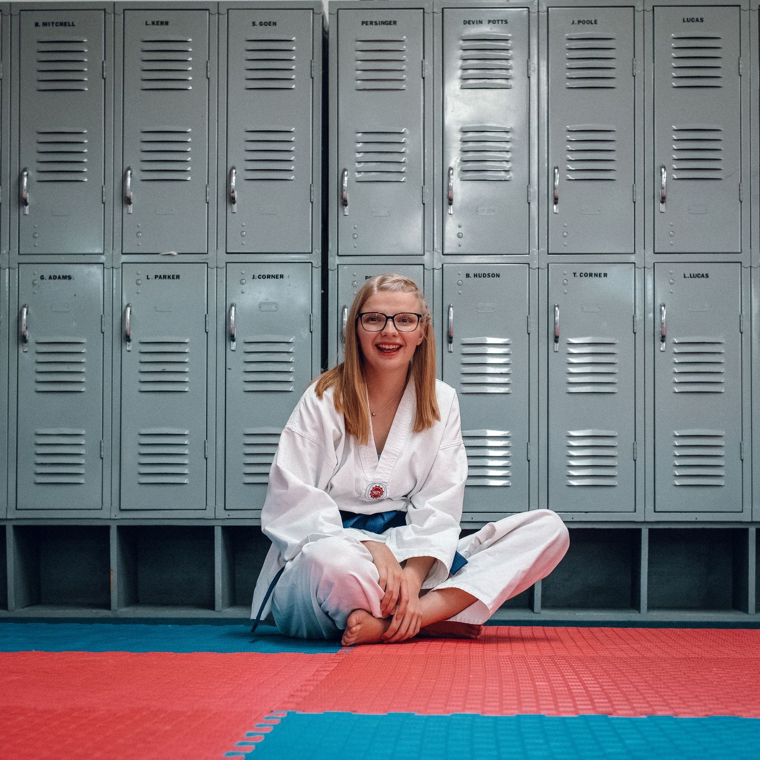 Smiling female martial artist in white gi with blue belt sitting on mat before gym lockers.