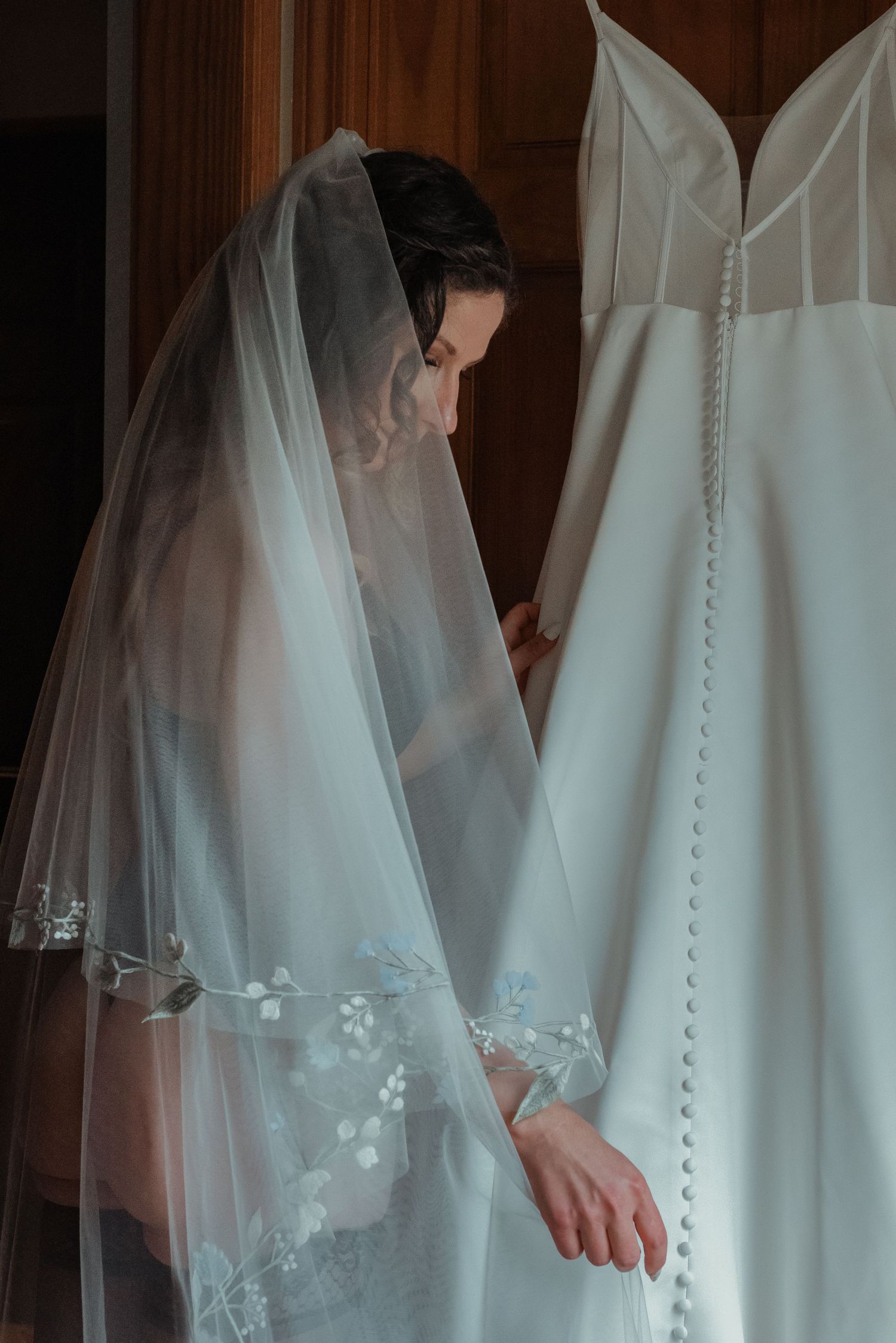 Bride wearing elegant lace veil stands beside her wedding dress in soft natural light.