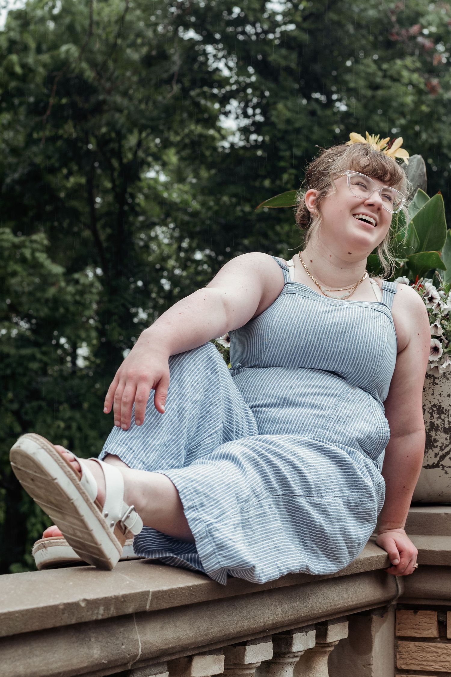 Young woman in striped sundress sitting on stone railing laughing outdoors.
