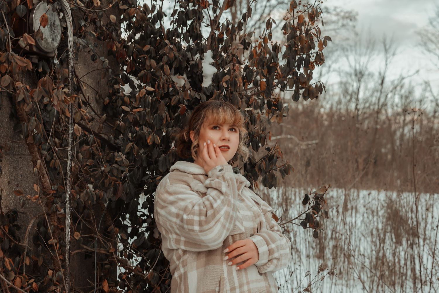 Young woman in plaid jacket standing by autumn foliage in a snowy winter landscape.