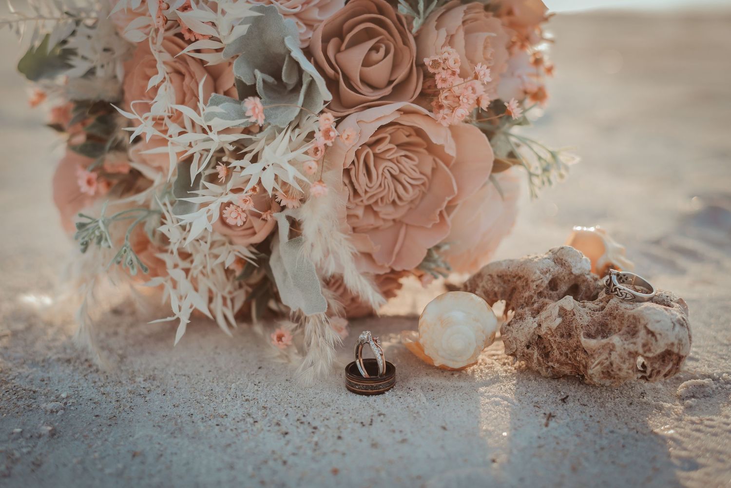 Romantic blush and peach bridal bouquet with roses and feathers beside wedding rings on sandy beach.