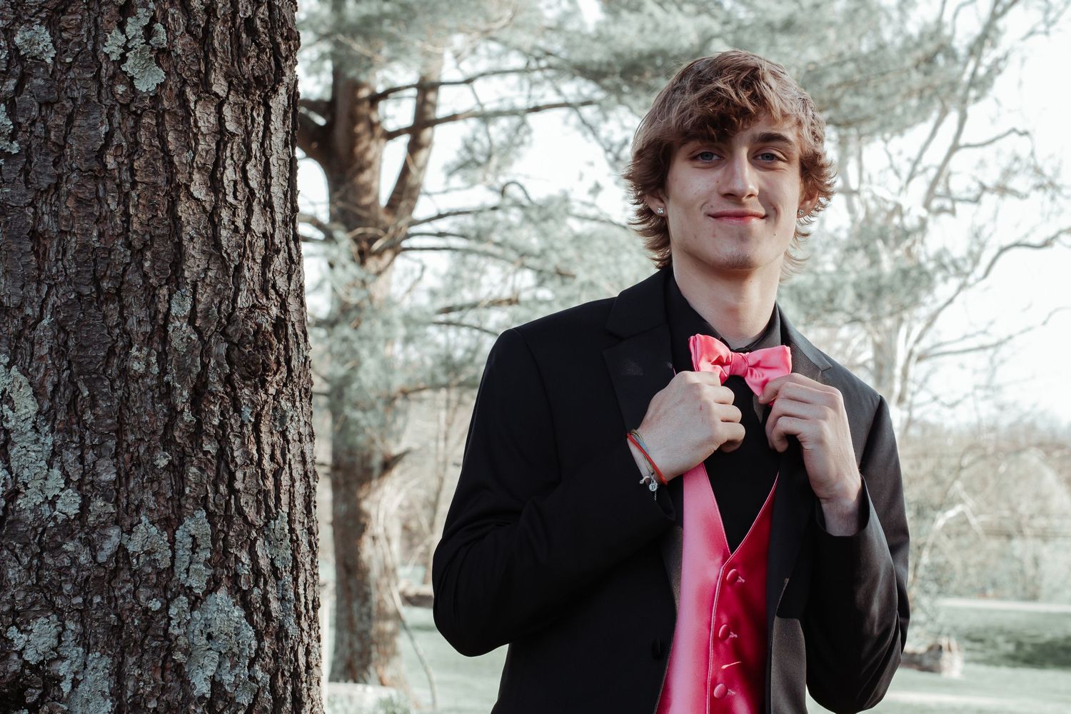 Young man in black tuxedo adjusts pink bow tie while standing by tree before prom.