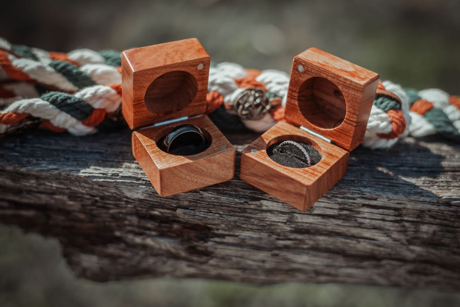 Two wooden ring boxes with wedding bands displayed on a rustic wood surface with a braided rope.