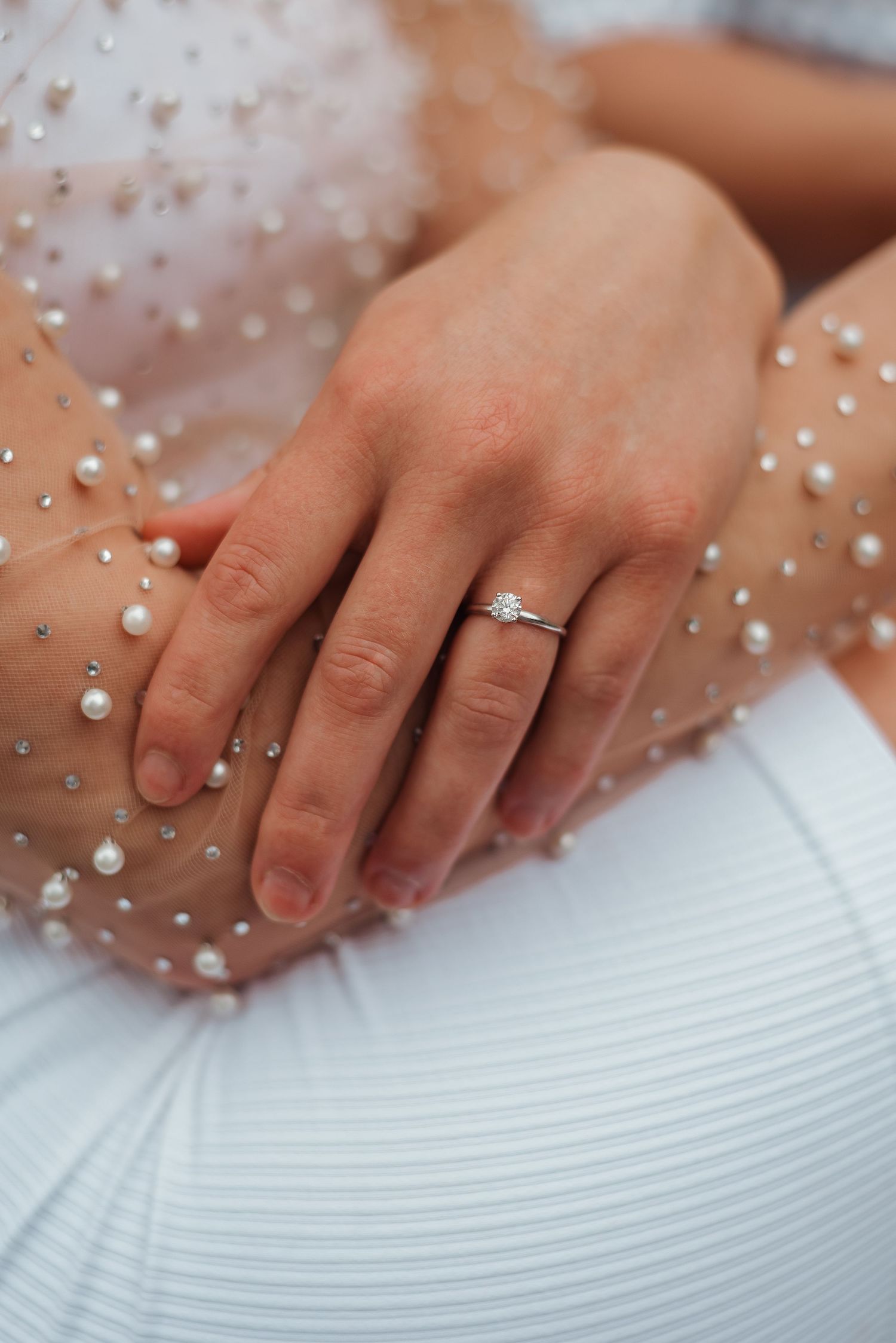 Close-up of an engagement ring with diamond solitaire on a woman's hand over white fabric.