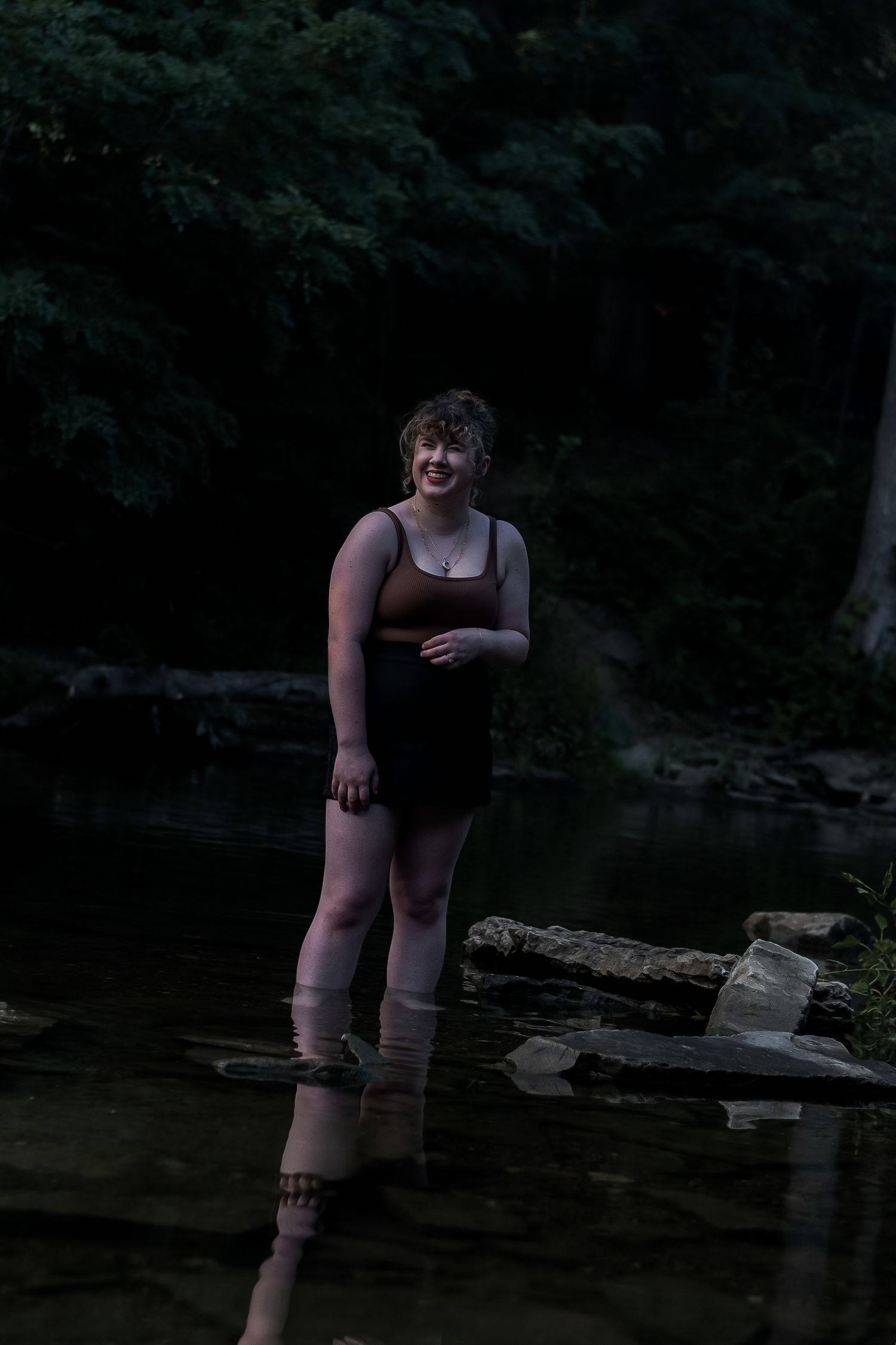 Young woman standing in shallow creek water smiling in dark forest setting.