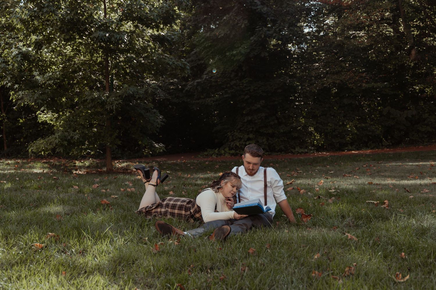 A couple relaxes on grass in a sunny park as the man reads aloud from a book.