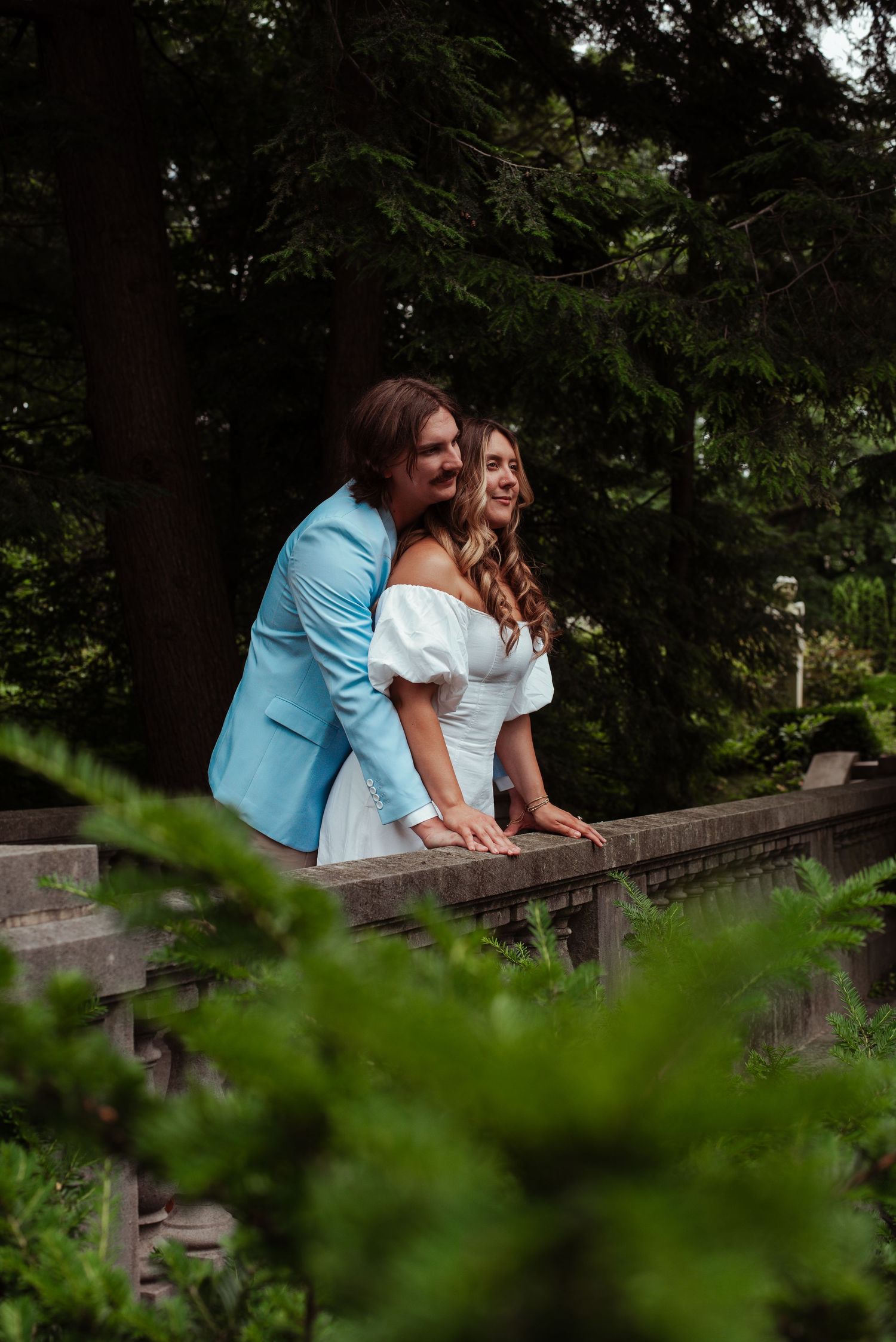 A couple in wedding attire smiles on a wooden bridge surrounded by lush green trees and ferns.