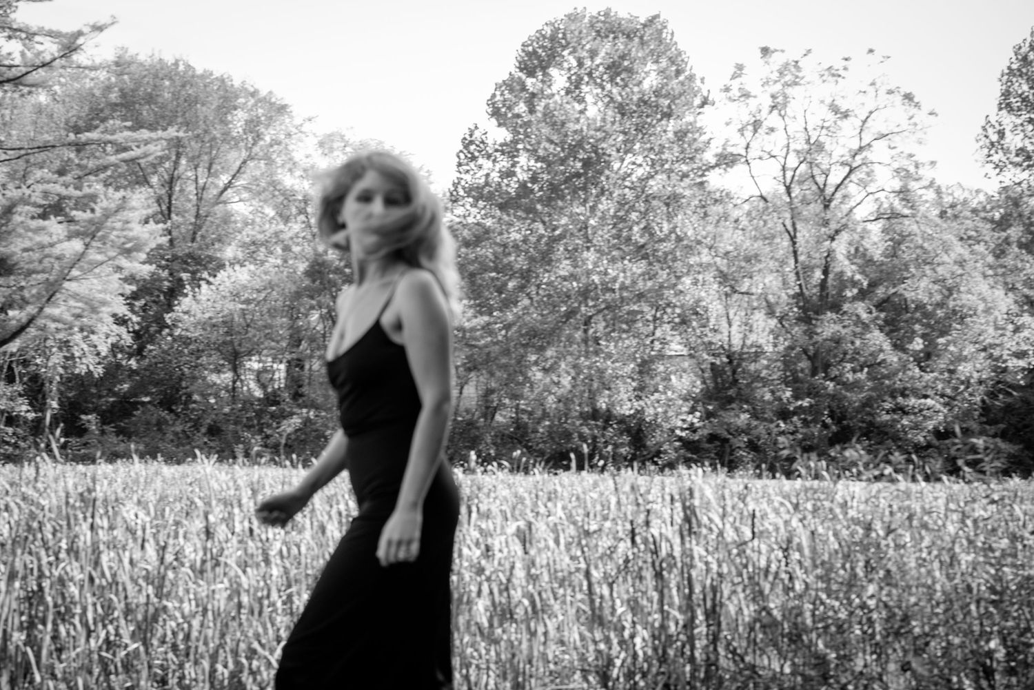 Black and white photo of blonde woman in black dress walking through a wild grass field.