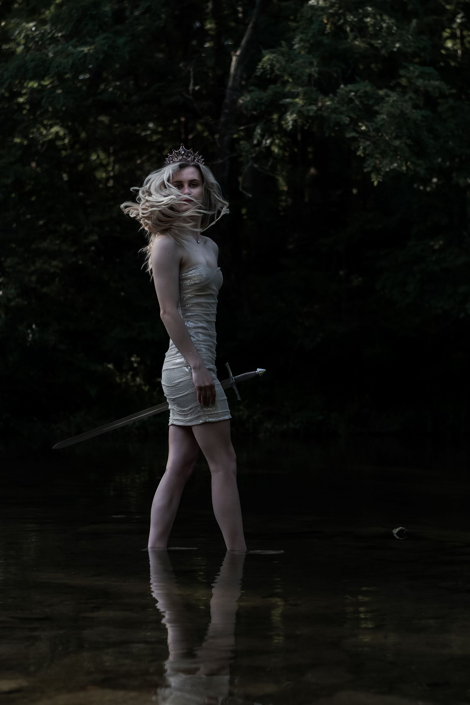 Blonde woman in white dress and crown stands in dark forest water holding a sword.
