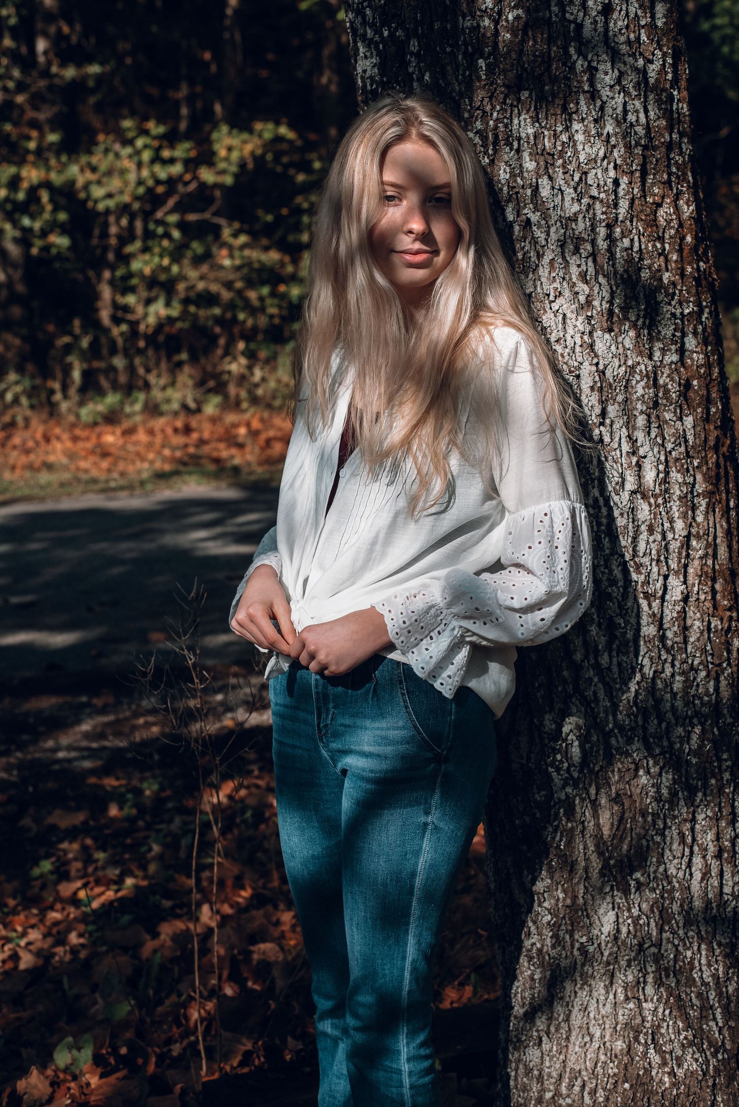 Blonde woman in white eyelet blouse leaning against tree trunk in autumn forest setting.