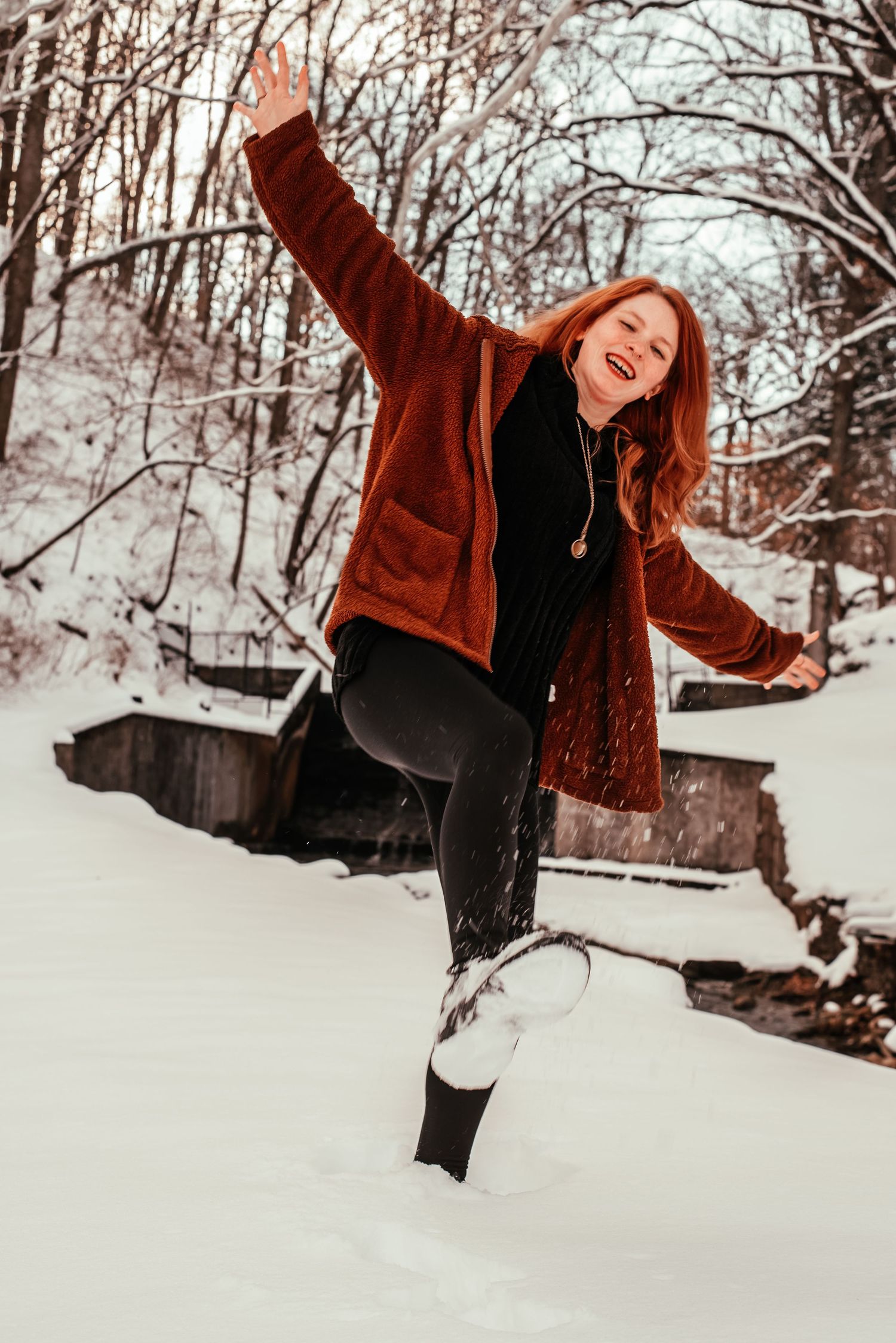 Joyful redhead woman in rust coat laughs and kicks snow in a snowy winter forest setting.