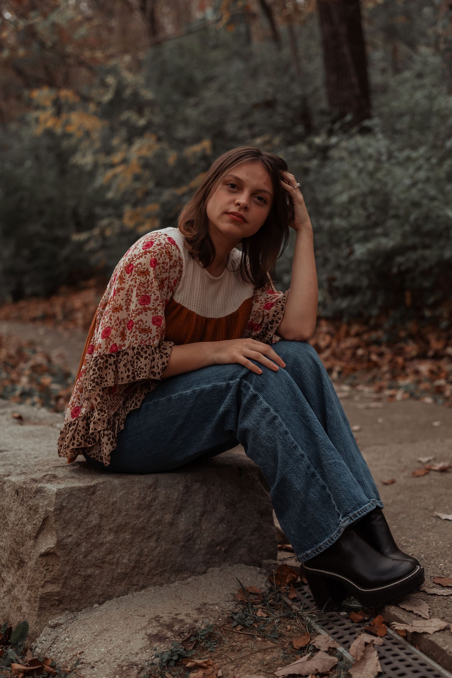 A young woman in a floral top and wide-leg jeans sits on a stone in an autumn forest.