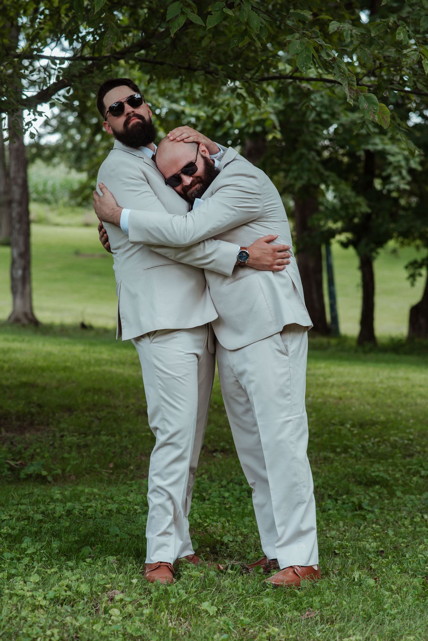 Two groomsmen in matching beige suits share a warm hug outdoors under green trees.