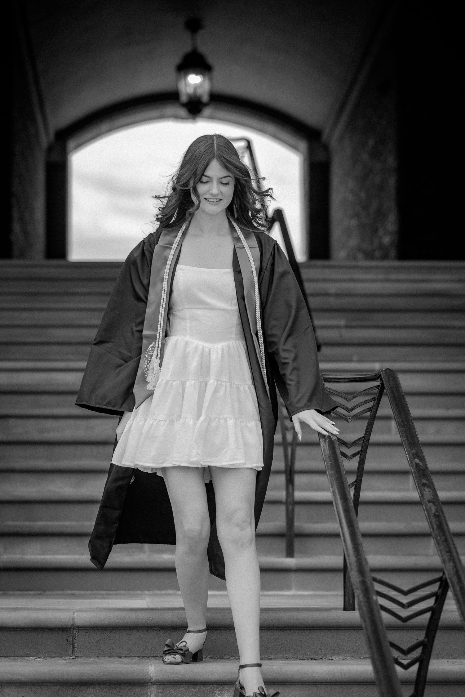 Black and white photo of a graduate in white dress descending ornate stone steps holding her gown.