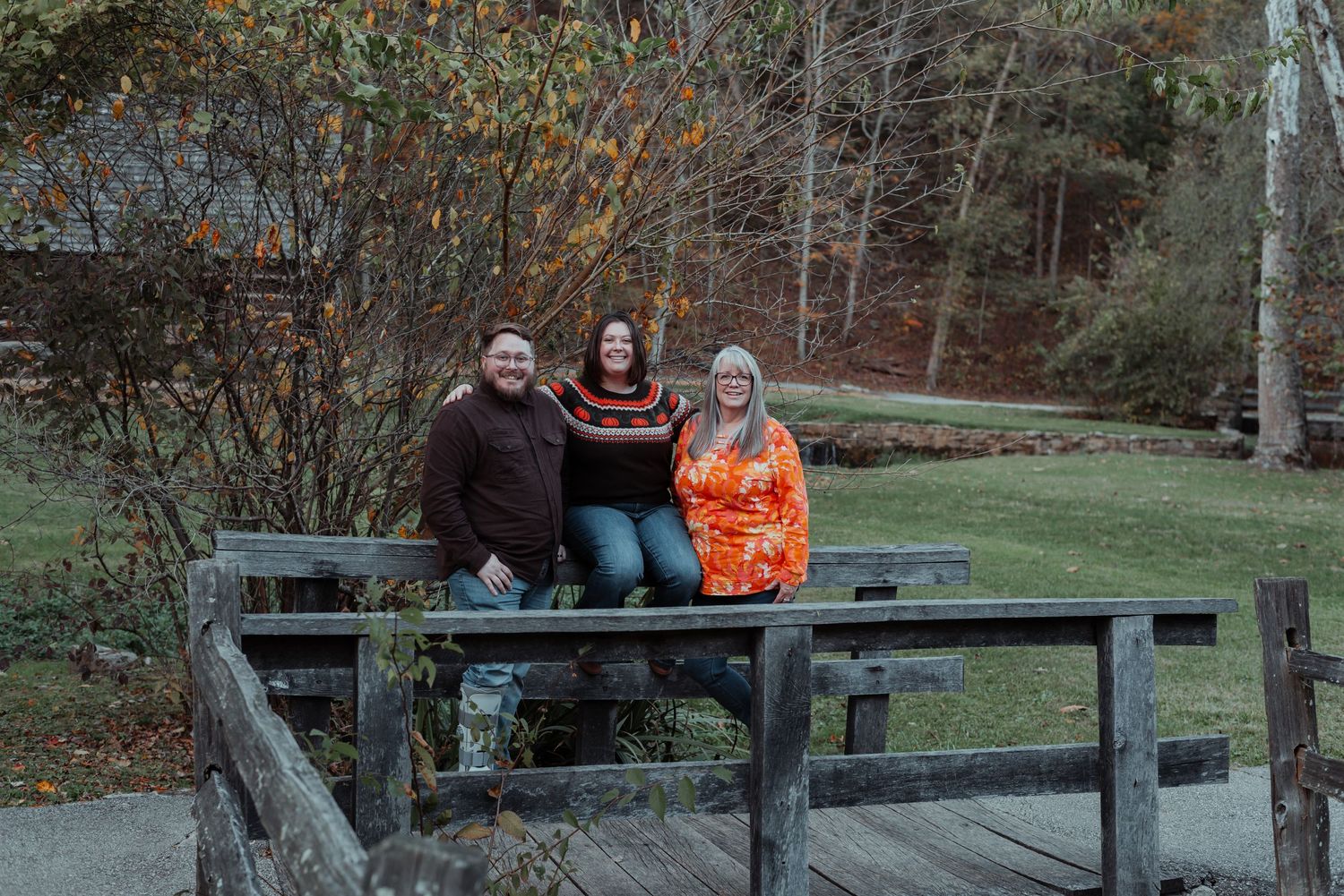 Three people smiling together on a wooden bridge amid colorful fall trees.