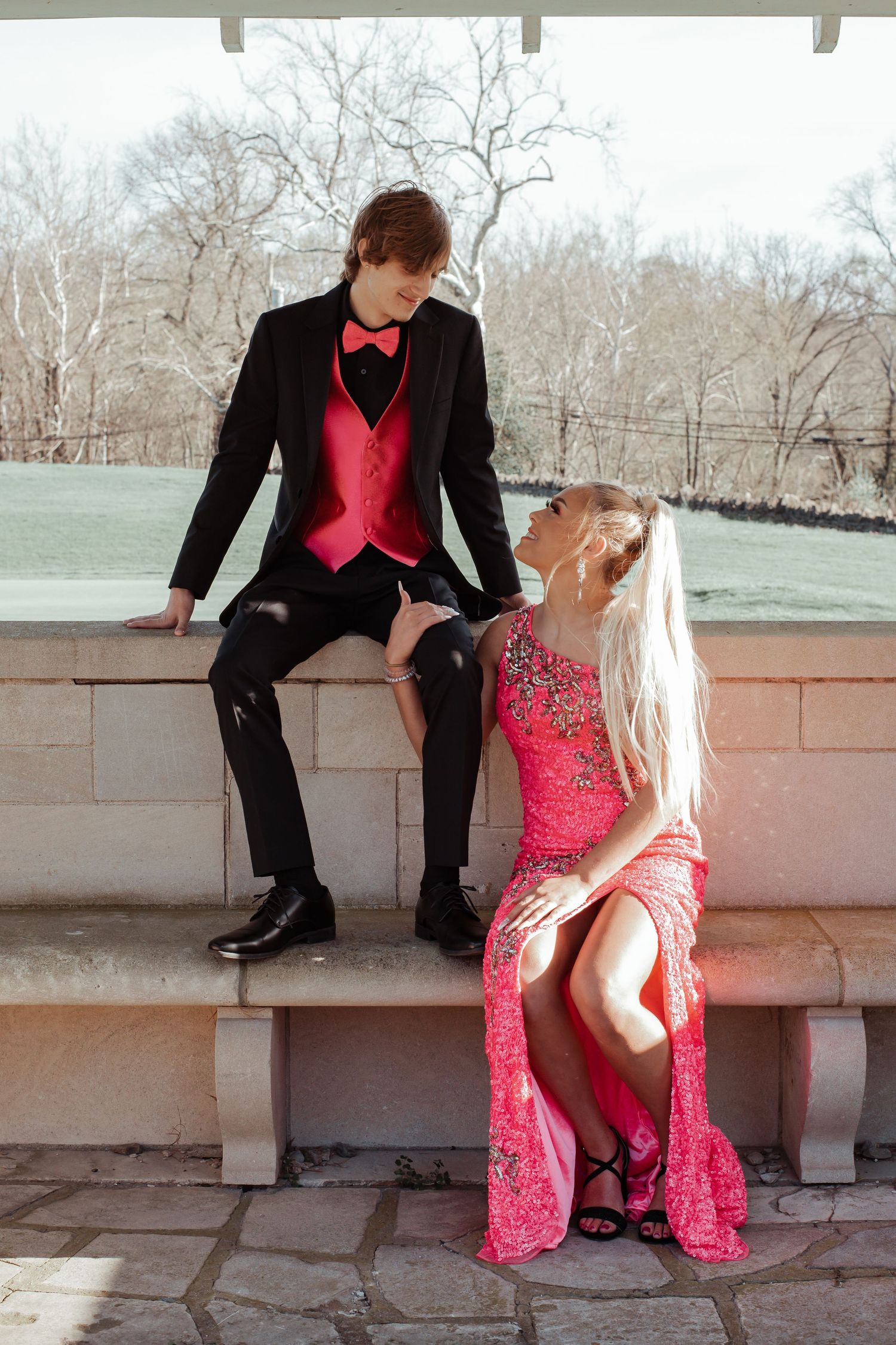Prom couple poses on stone ledge, boy in black tuxedo with red vest, girl in pink beaded gown.