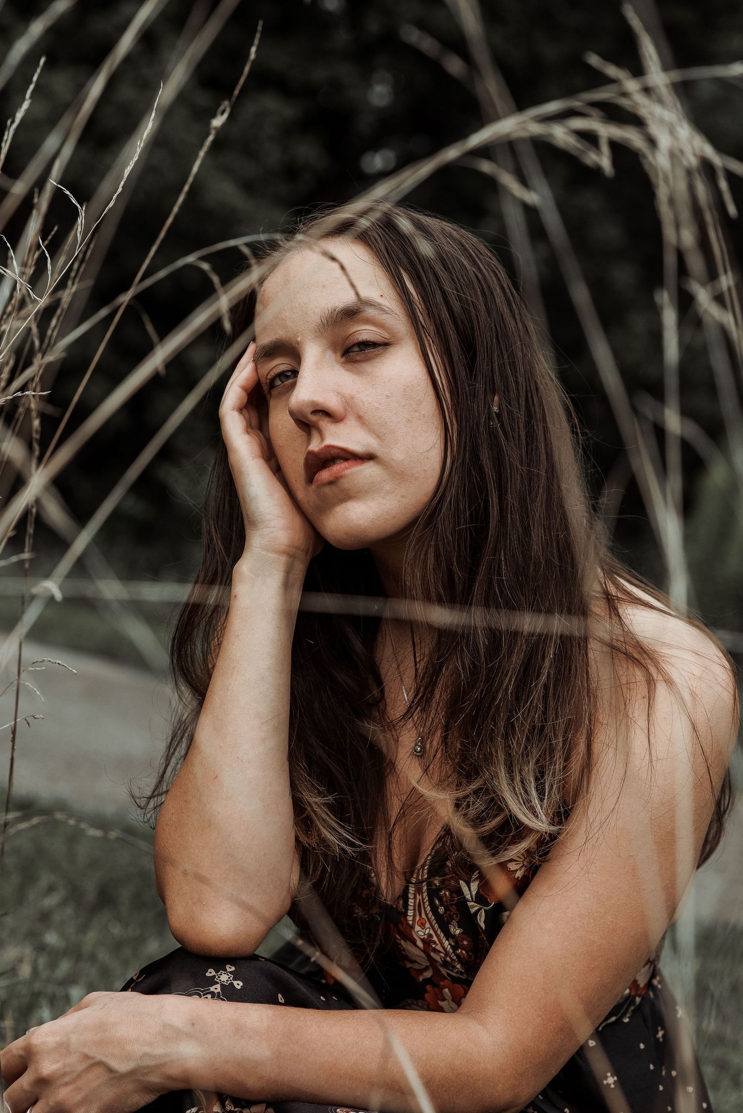 Brunette woman resting hand on face while sitting among tall dry grasses.