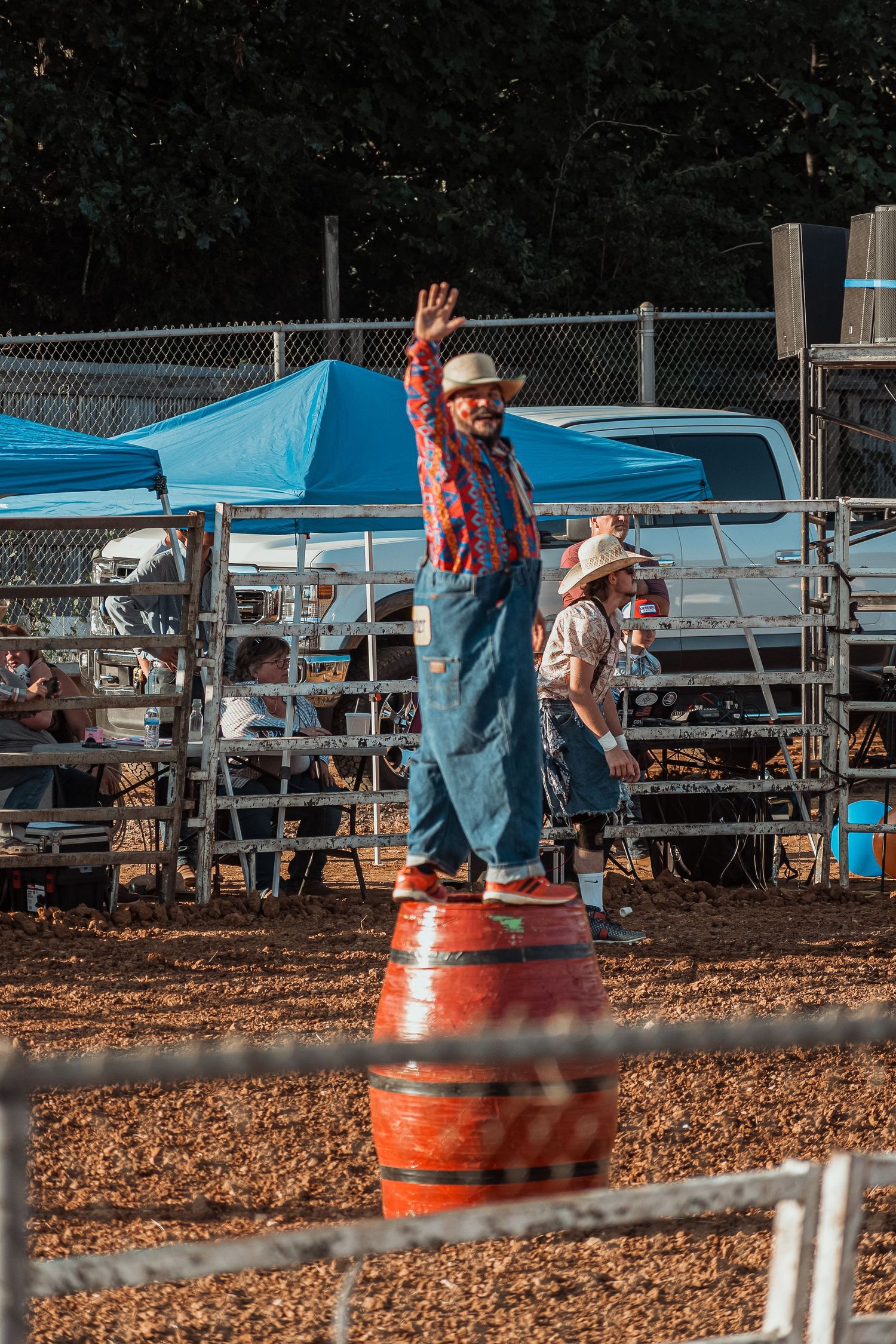 A rodeo clown in overalls and a hat waves while standing on a barrel at a rodeo event.