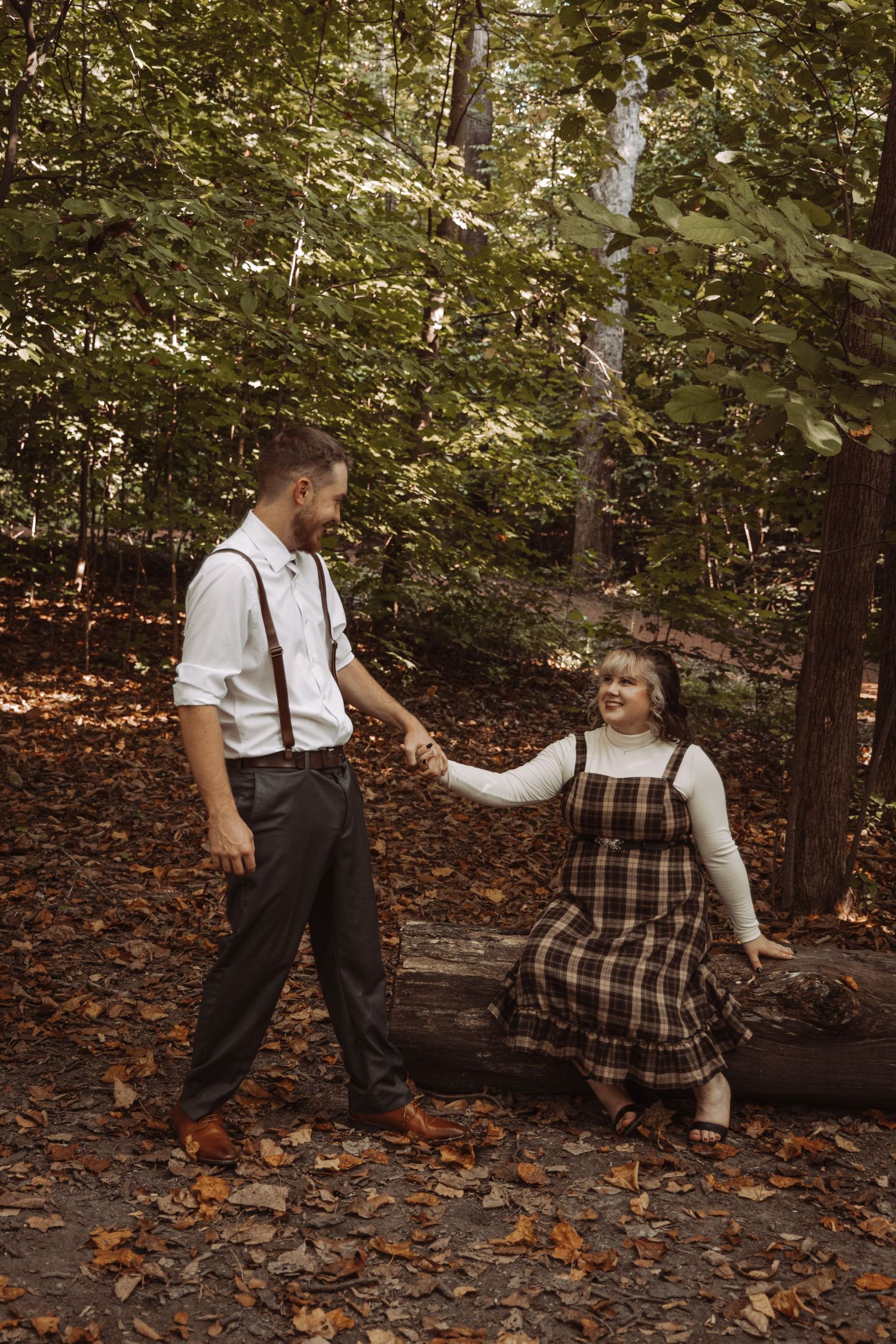 Couple holding hands in an autumn forest, woman in plaid pinafore dress sitting on a log, man in suspenders.