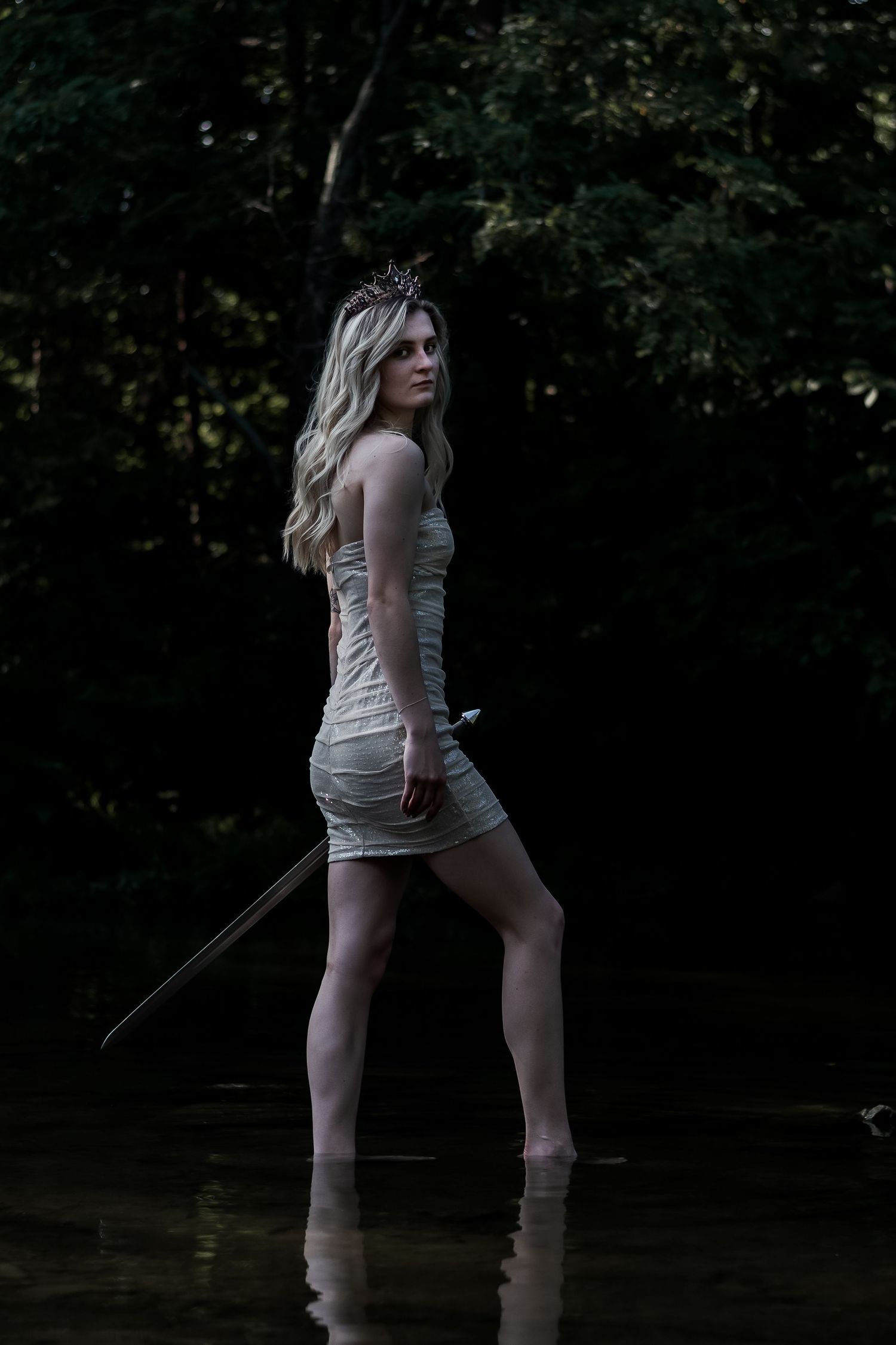 Mysterious blonde warrior queen in white dress wades through dark forest water with sword.