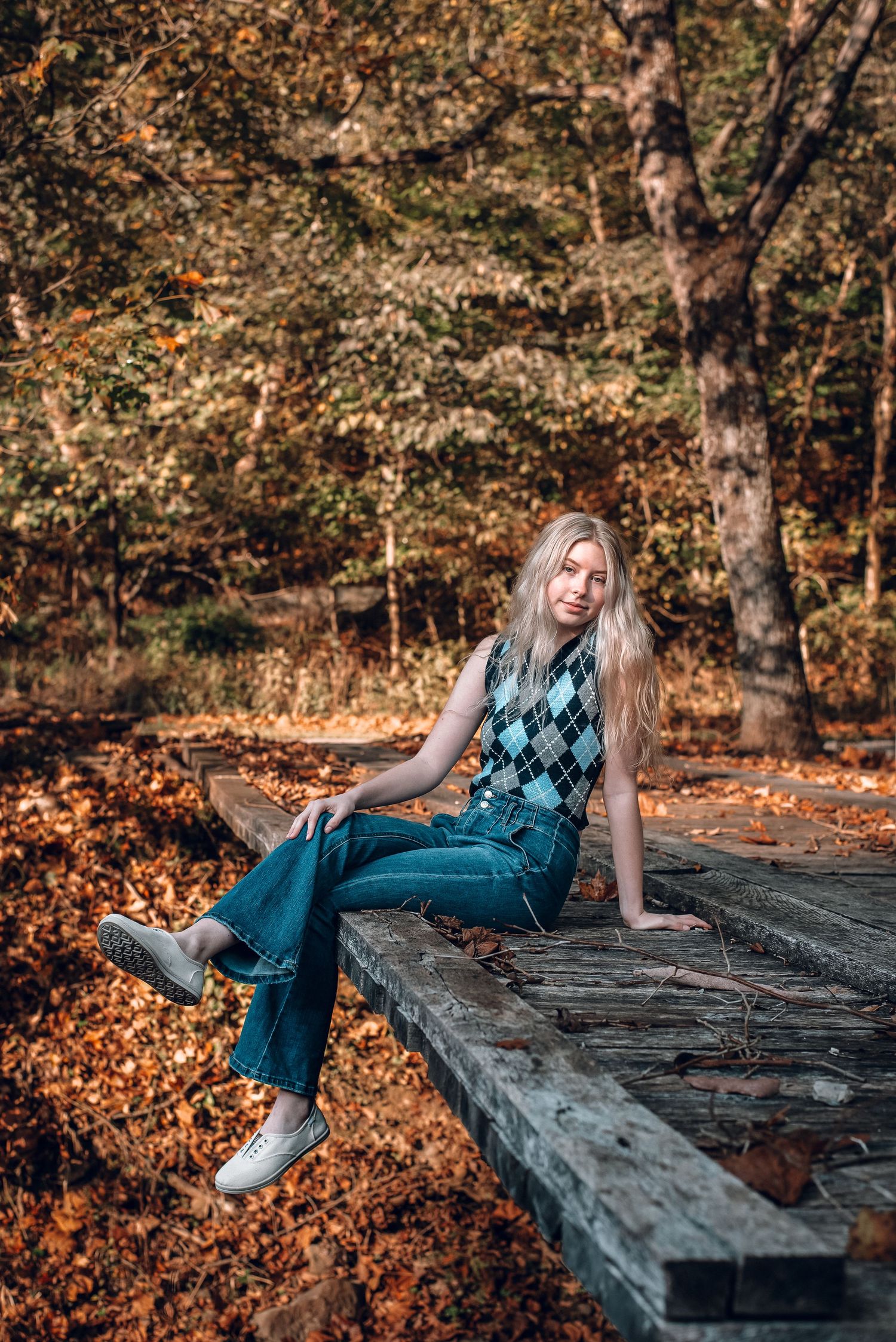 Blonde woman in argyle vest and flared jeans sitting on wooden platform in autumn forest.