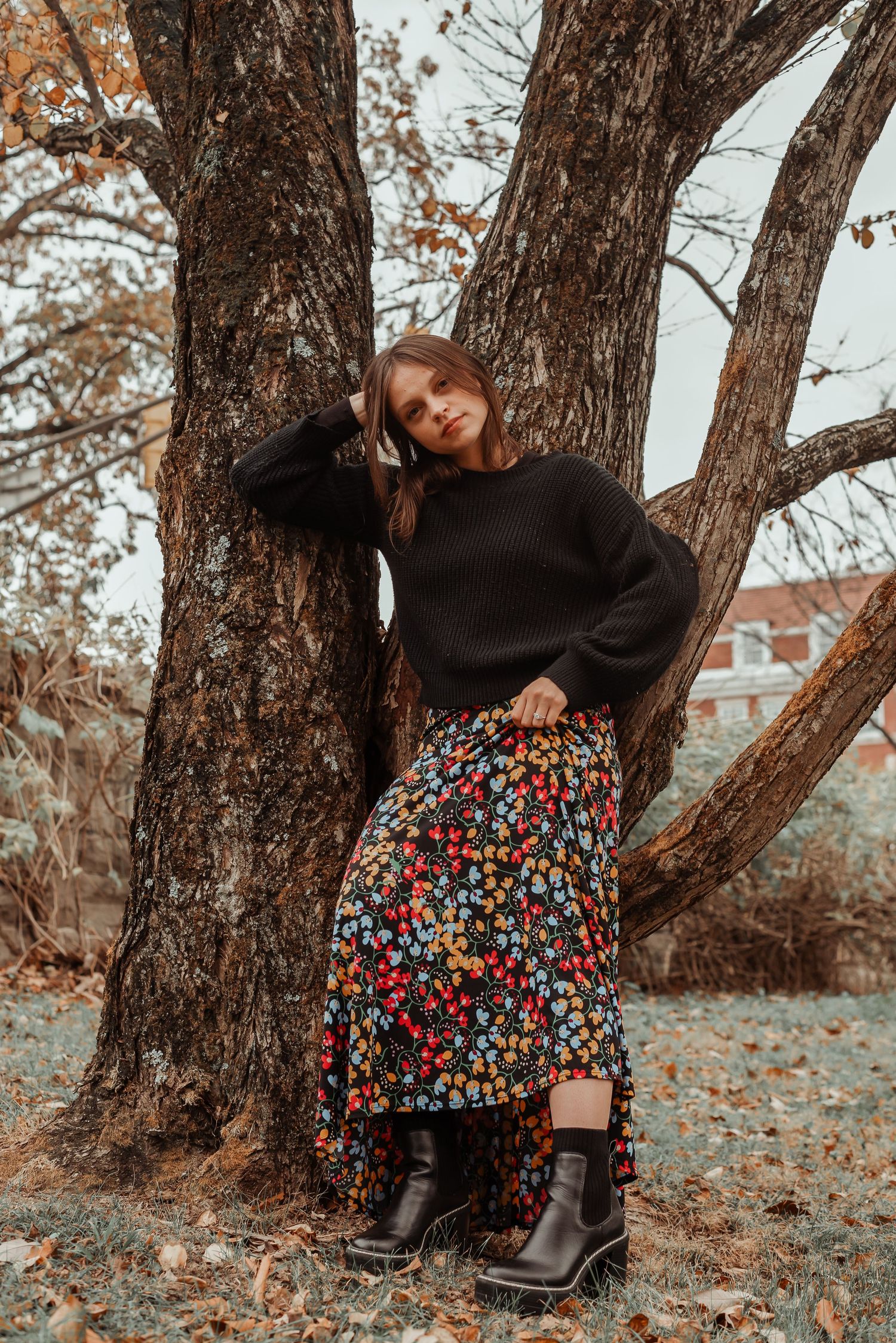 Young woman in black sweater and colorful floral skirt leans against tree in autumn park.