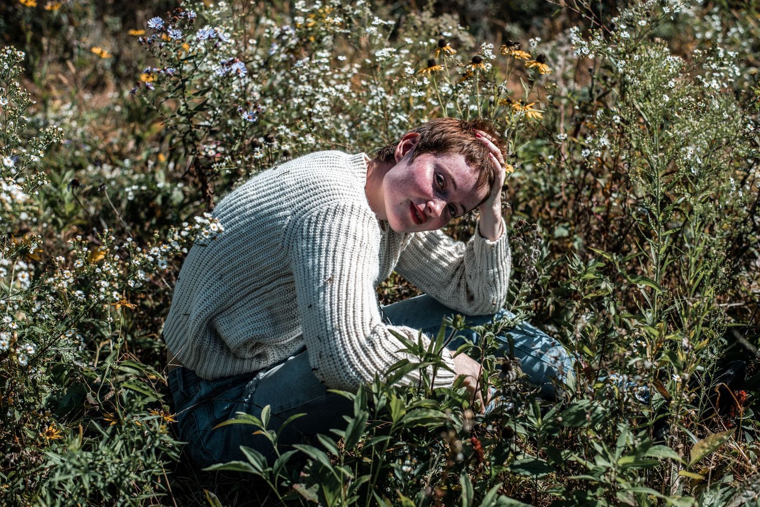 Person in cream sweater leaning among wildflowers in a sunny meadow.