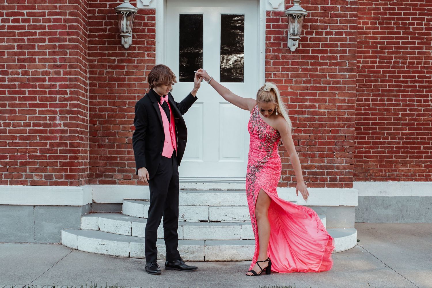 Prom couple dances outside brick building, boy in black suit with pink tie, girl in pink gown.