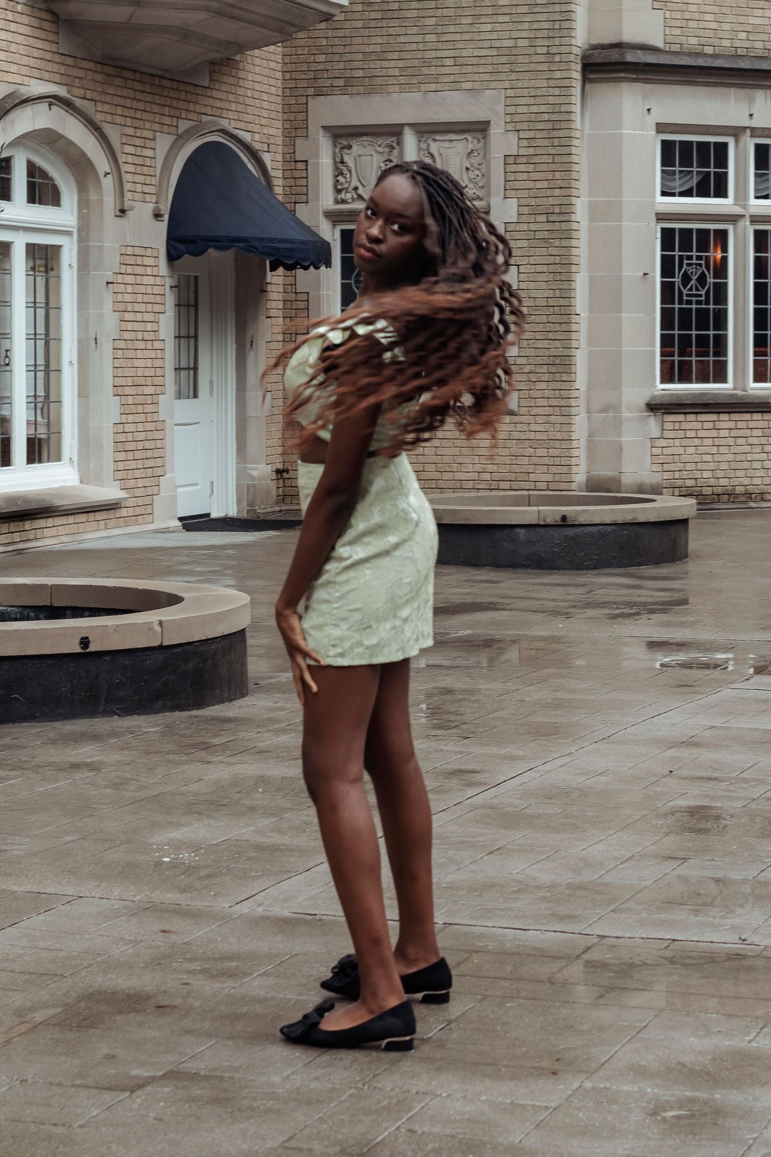 Young woman in green mini skirt set tossing hair while posing on wet courtyard pavement.