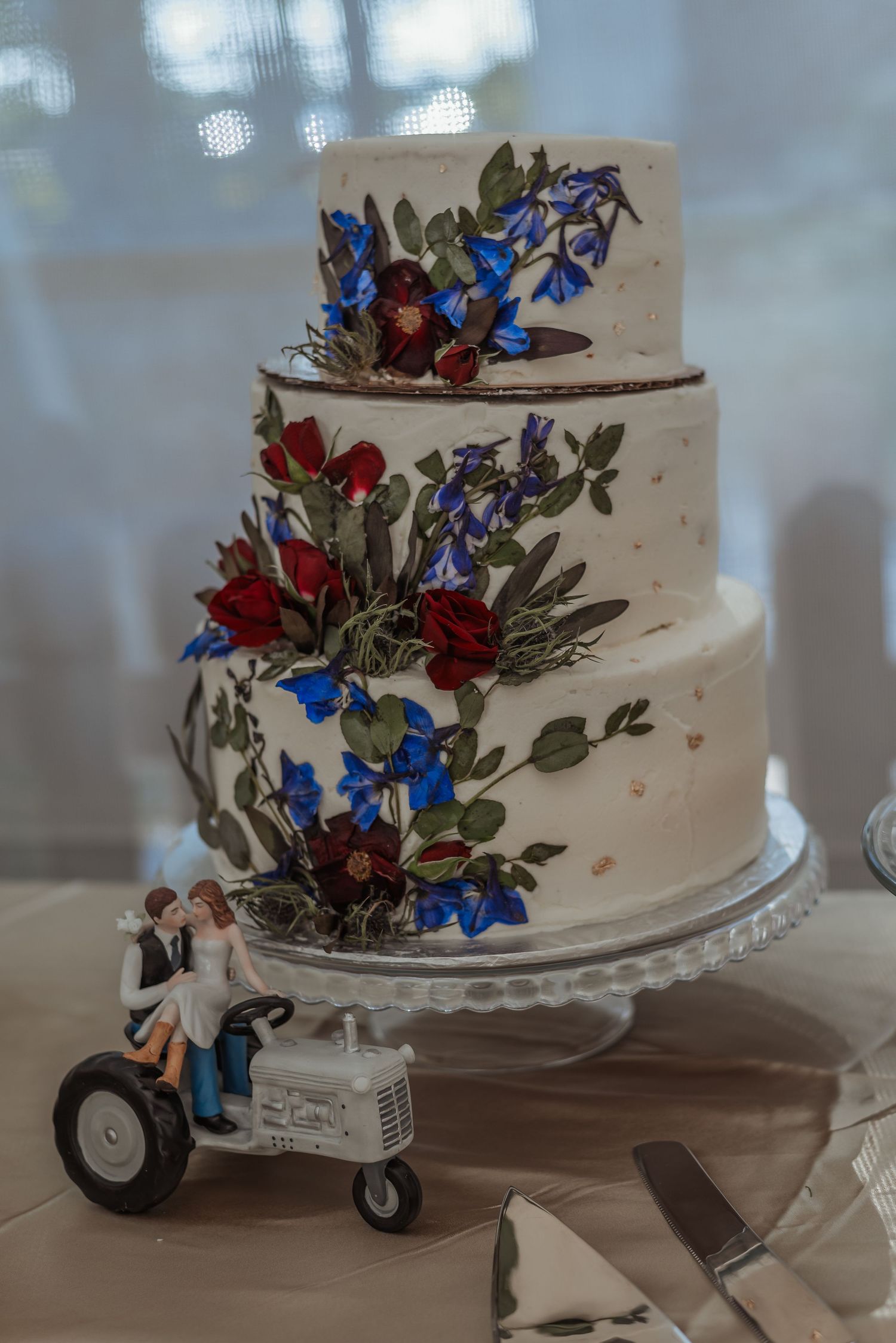 Three-tier white wedding cake decorated with red roses and blue flowers with tractor topper.