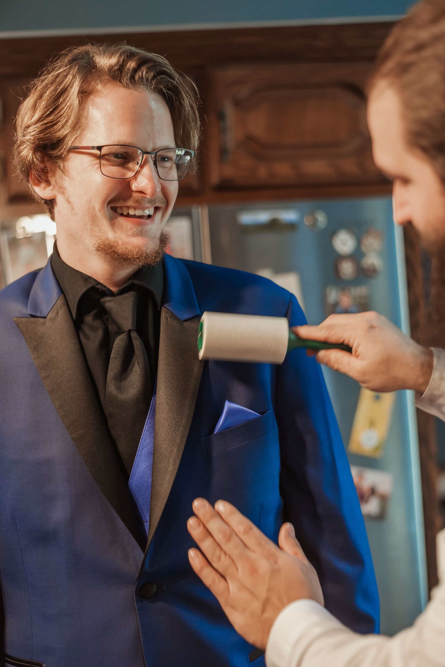 Smiling groom in royal blue tuxedo being lint-rolled by groomsman while getting ready indoors.