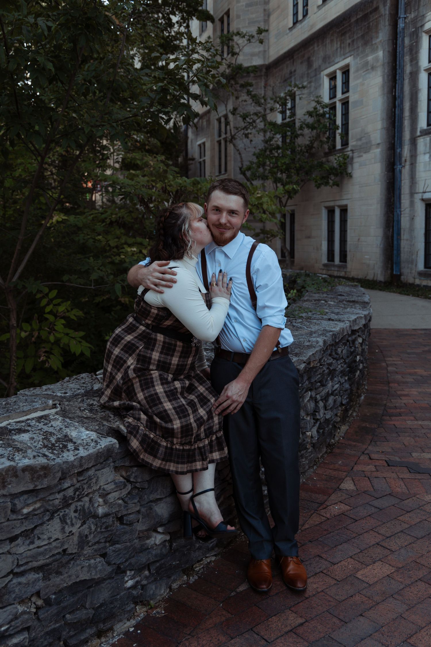 A smiling couple embraces on a stone ledge outside a historic stone building at dusk.