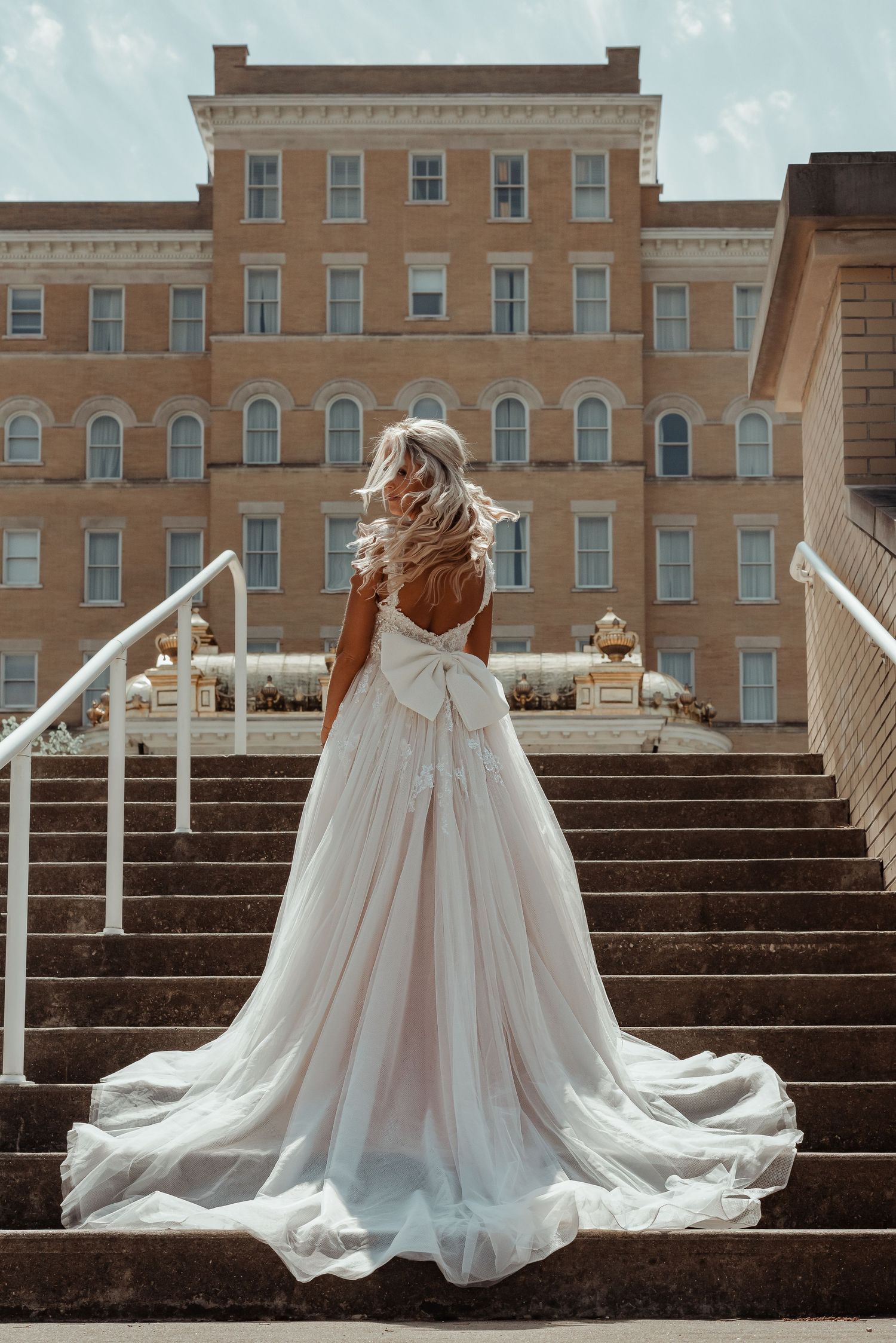 Bride in a stunning backless white ball gown with bow standing on stone steps before a grand building.
