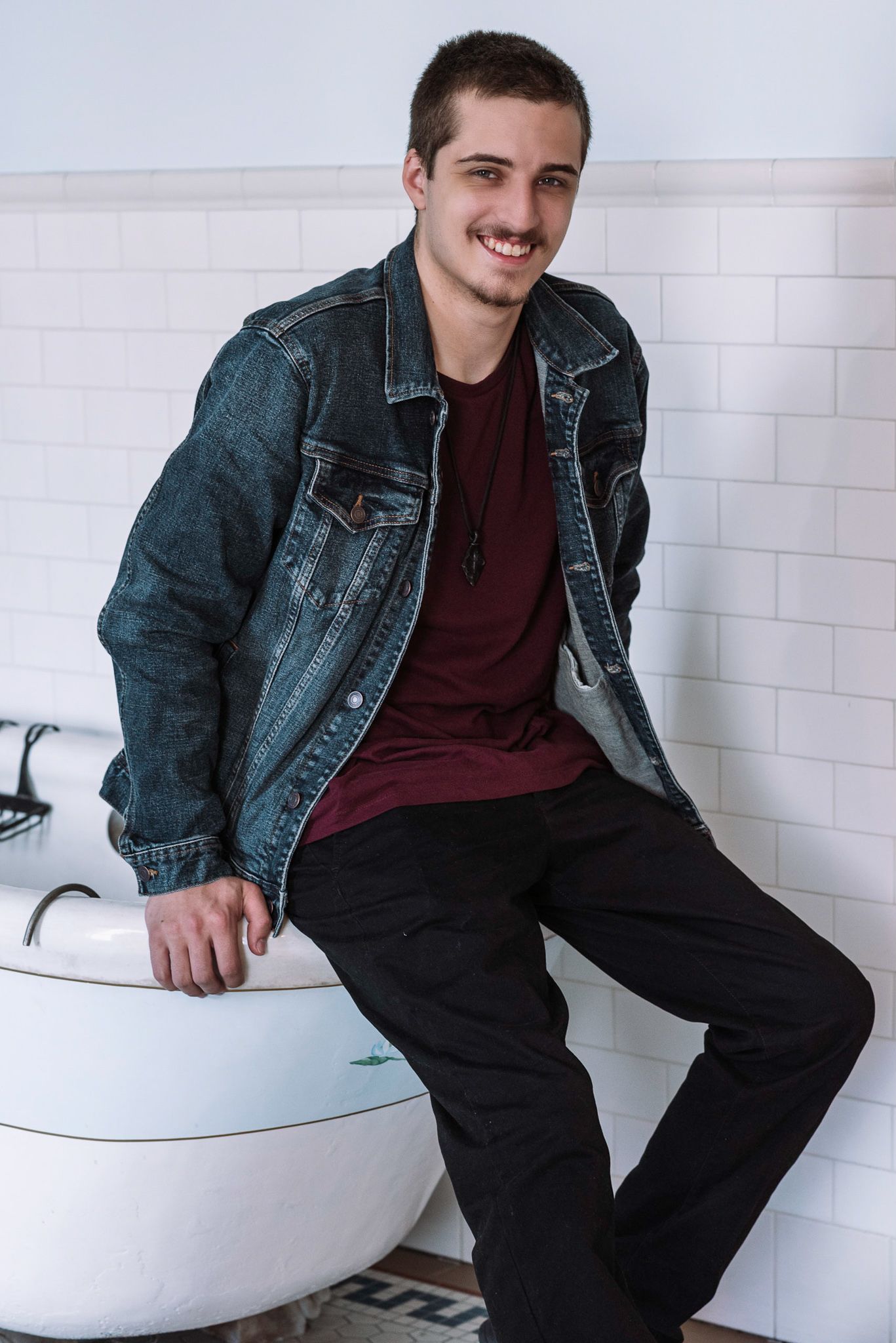Smiling young man in denim jacket sitting on bathtub edge indoors.