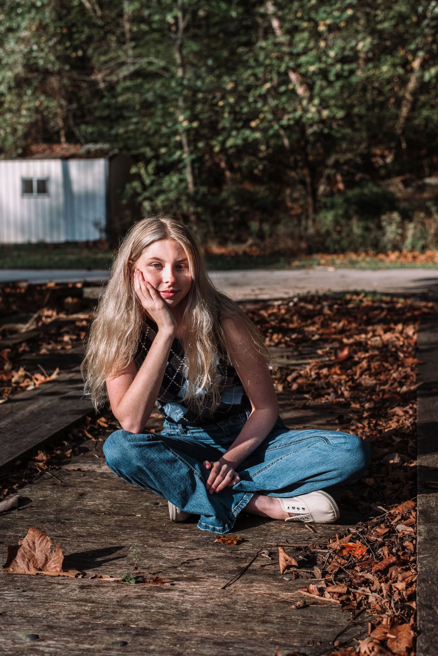 Blonde woman sitting cross-legged on wooden deck surrounded by autumn leaves in forest setting.