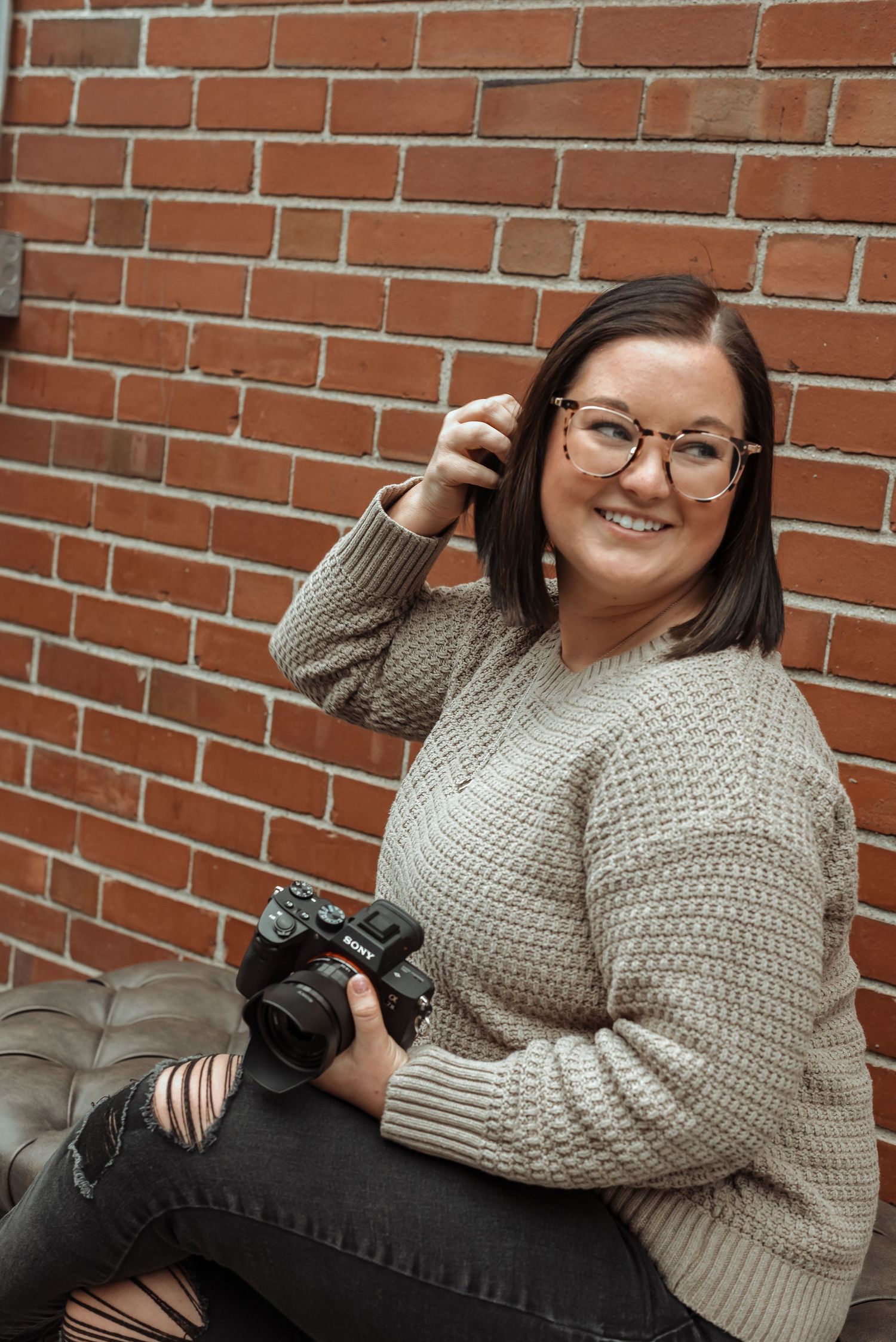 Smiling brunette woman holding a Sony camera, seated against a red brick wall in a beige sweater.