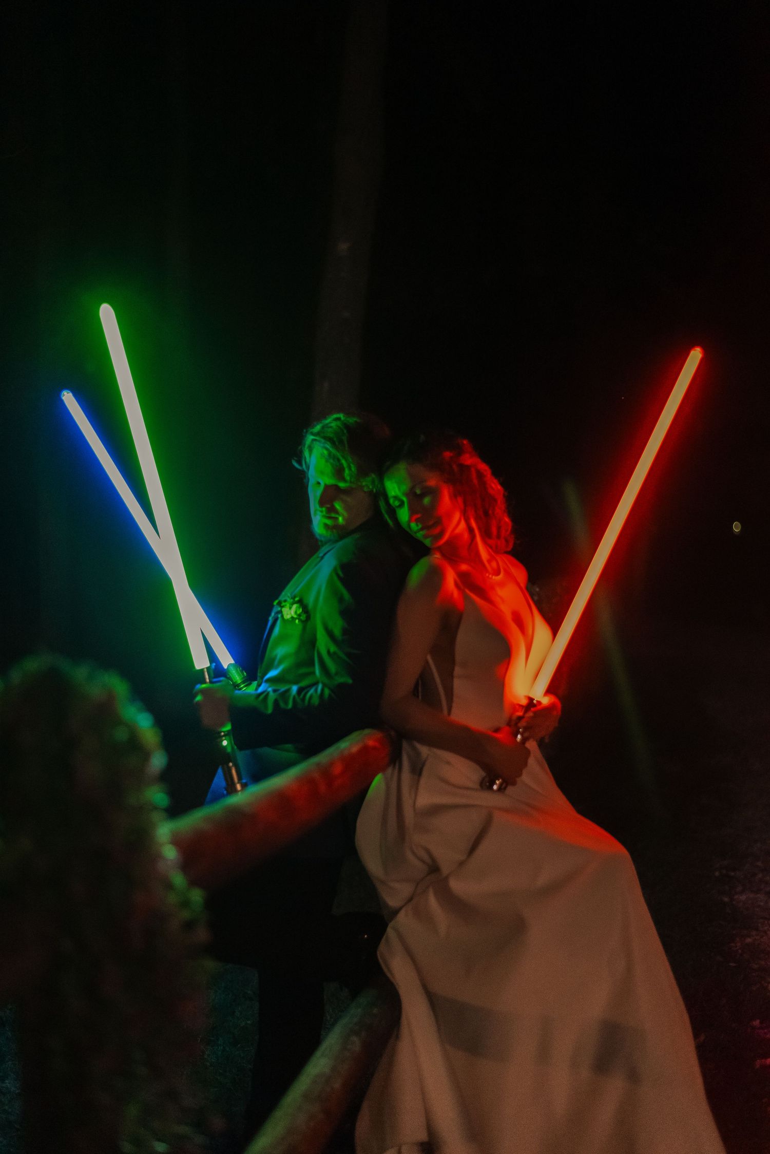 Bride and groom posing dramatically with colorful lightsabers in dark outdoor Star Wars wedding.