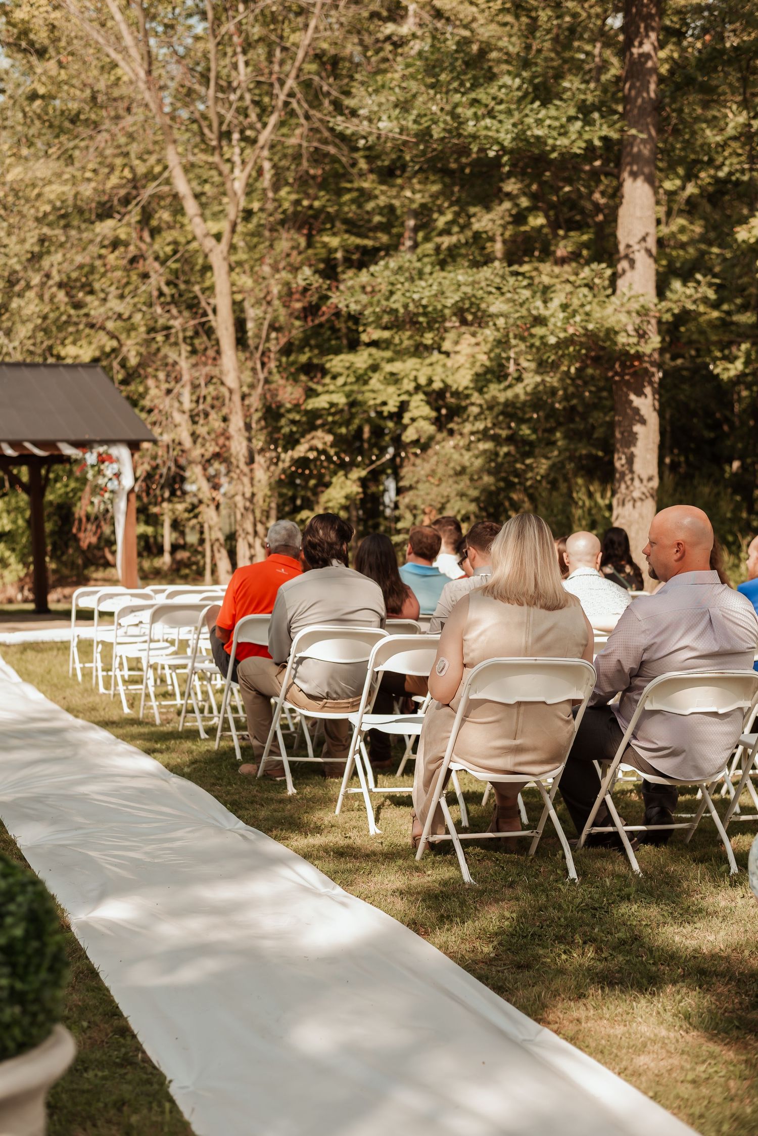 Guests seated in white folding chairs at an outdoor wedding ceremony surrounded by lush green trees.