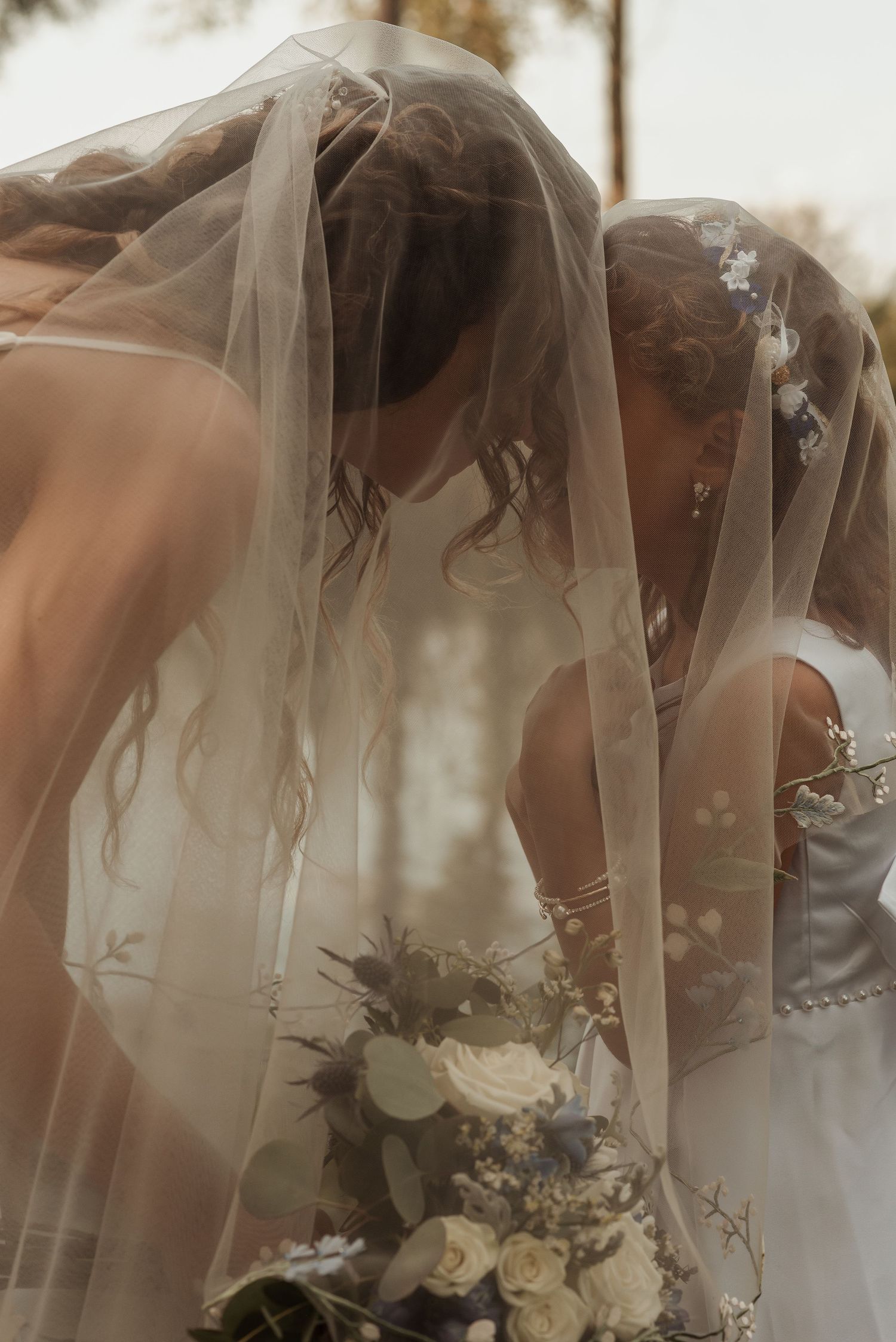 A bride and bridesmaid share an intimate moment hidden beneath a flowing wedding veil.
