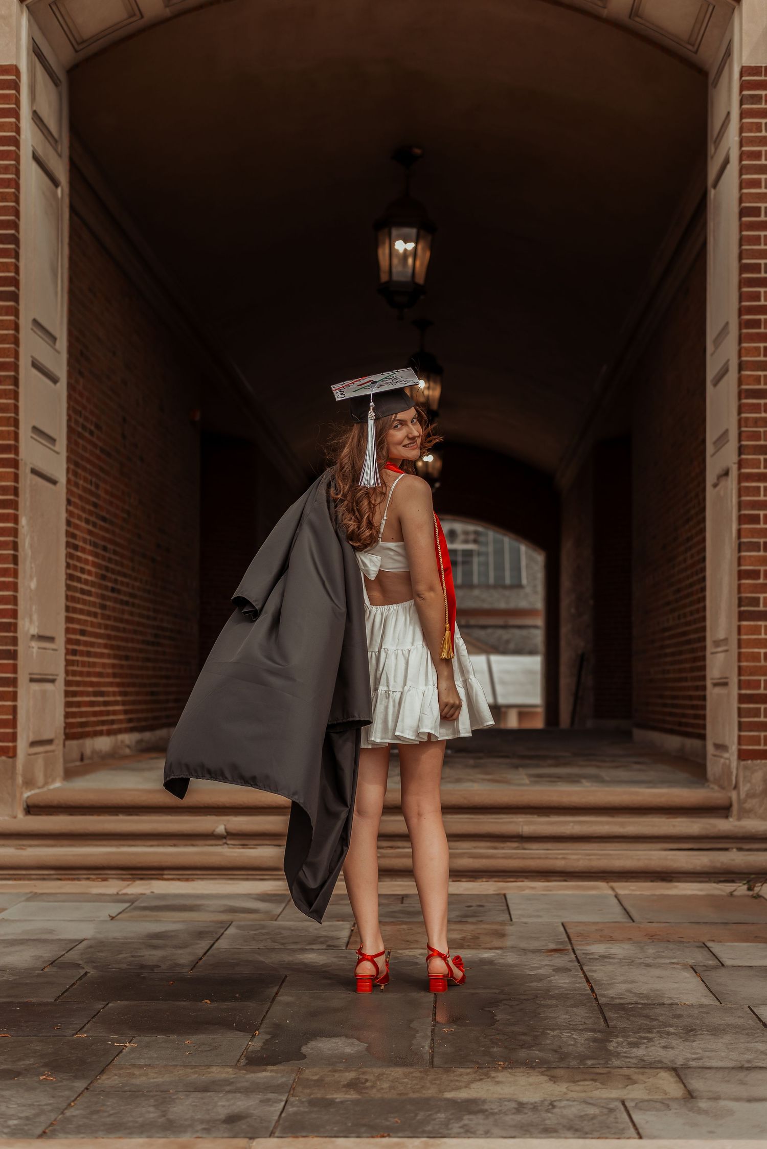 Graduate in white dress and red heels poses with black gown in arched brick hallway wearing mortarboard cap.