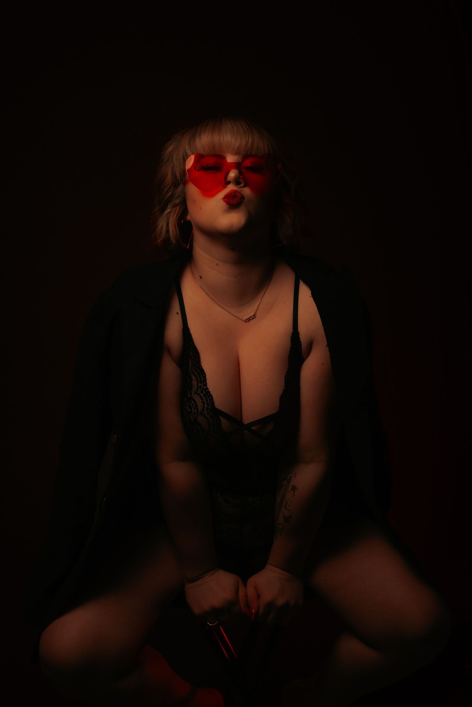 Woman in black lace bodysuit with red heart sunglasses in dark studio.